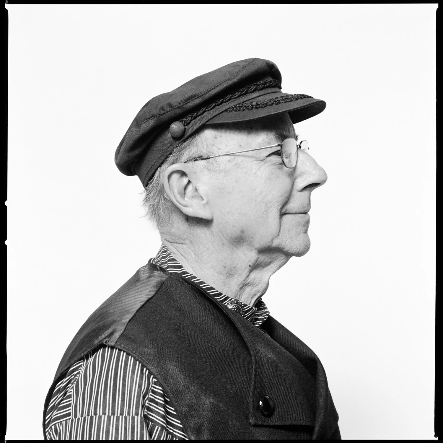 Black and white side profile of an elderly man wearing glasses, a striped shirt, and a dark cap with decorative braids, against a plain light background. Pella, Iowa Tulip Time Festival, Dutch Heritage