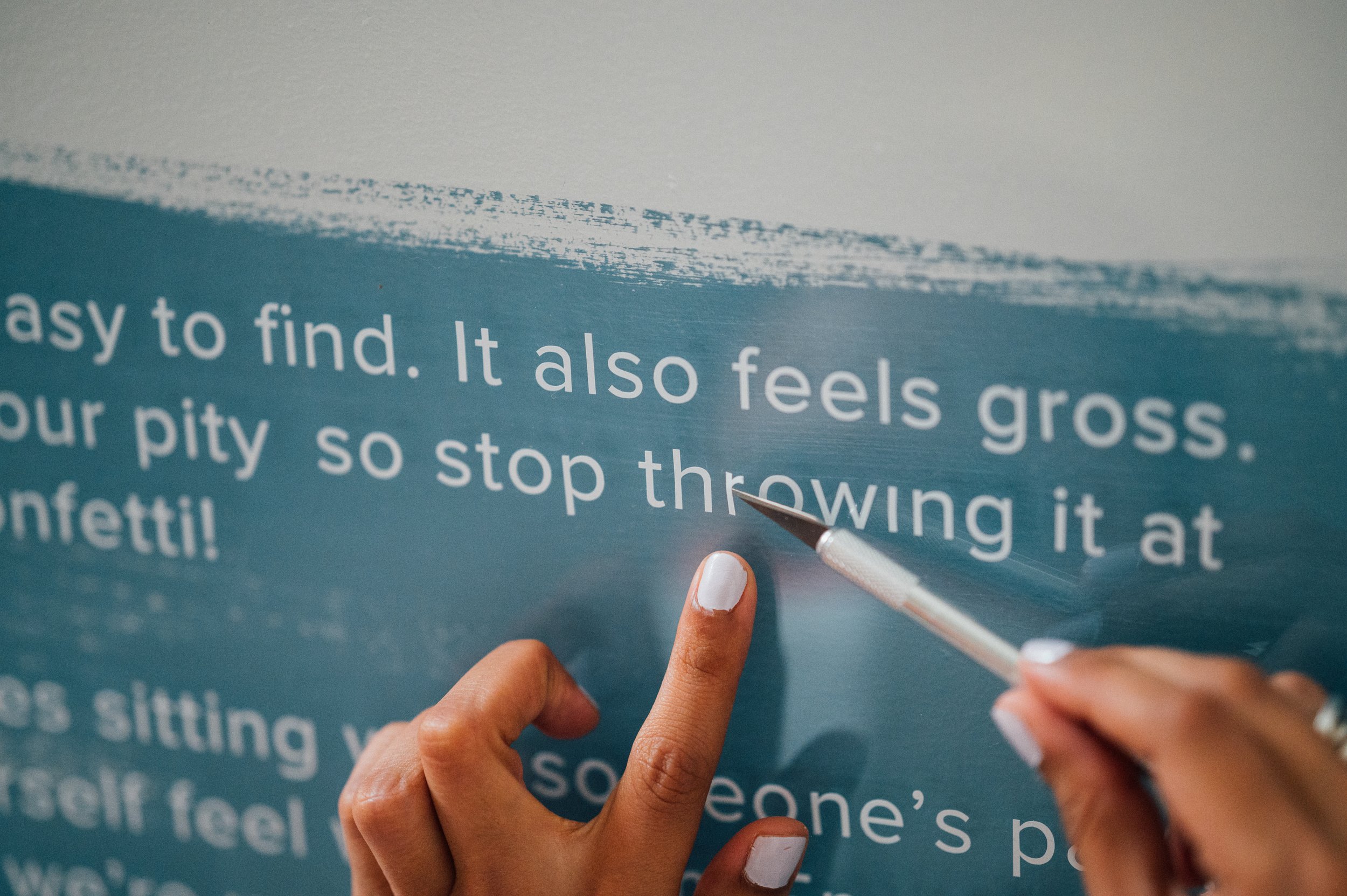 Close-up of a person painting white text on a blue wall with a small brush, focusing on their hand and painted words.