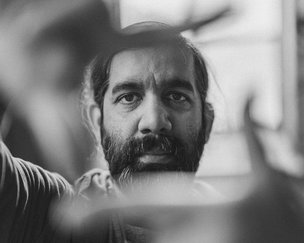 A black and white close-up of a man with a beard and mustache, looking directly at the camera, with blurred surroundings. Sk8 Liborius