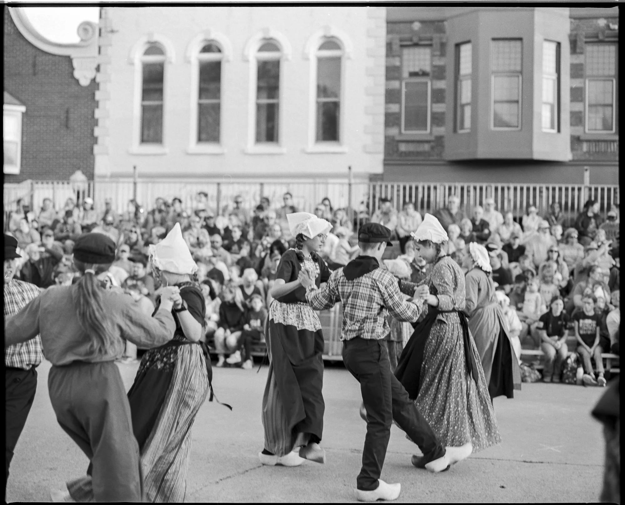 People dressed in traditional folk costumes dancing outdoors in front of a crowd of onlookers, with historic buildings in the background. Pella, Iowa Tulip Time Festival, Dutch Heritage