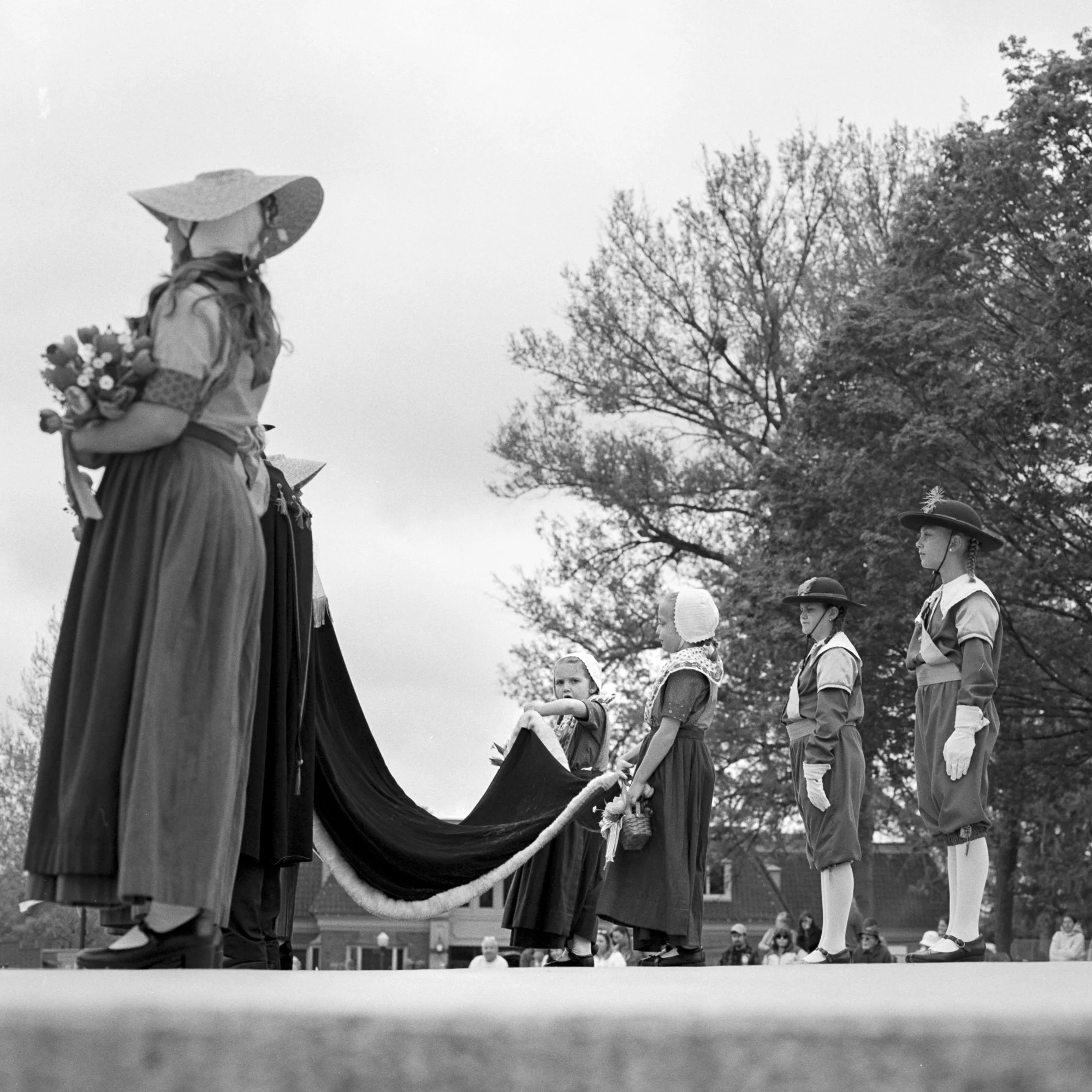 A black-and-white photo of children dressed in historic costumes, standing in a line outdoors with trees in the background, participating in a reenactment or celebration. Pella, Iowa Tulip Time Festival, Dutch Heritage