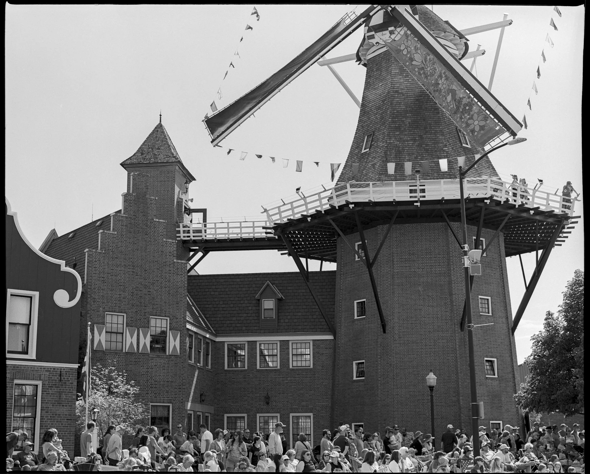A large crowd gathered outside a historic windmill at a theme park or festival, with many people sitting and standing, some wearing hats and sunglasses, under a cloudy sky. Pella, Iowa Tulip Time Festival, Dutch Heritage