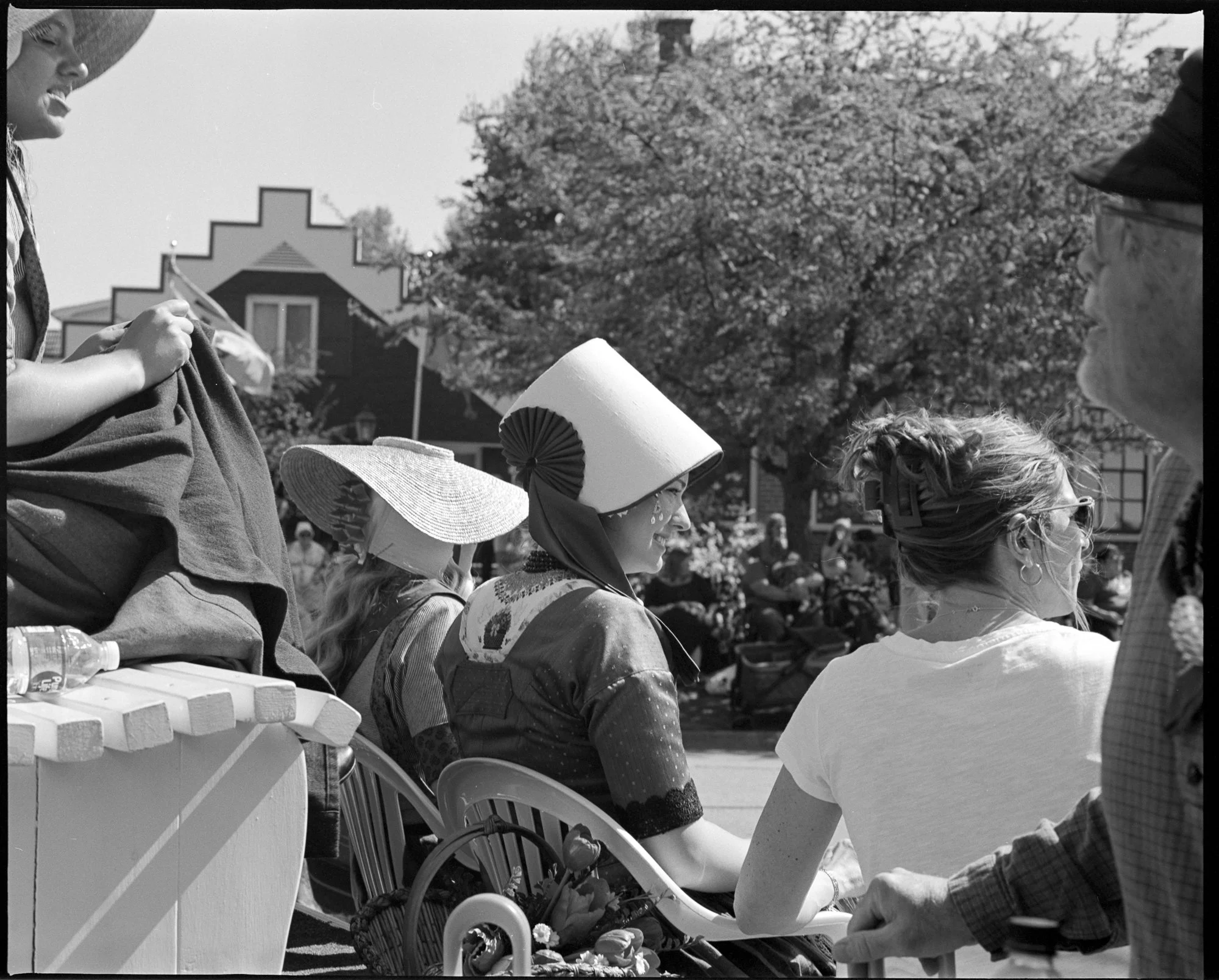 People sitting outdoors at a parade or festival, some wearing wide-brimmed hats and summery clothing, with a house and trees in the background. Pella, Iowa Tulip Time Festival, Dutch Heritage