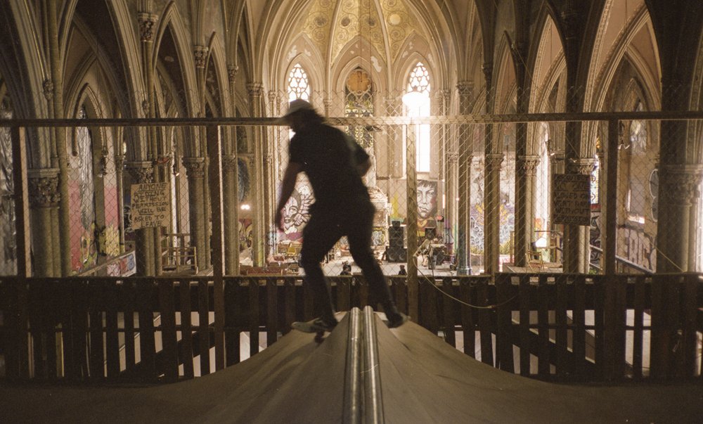 A person skateboarding on a ramp inside a large, gothic-style church with stained glass windows and graffiti on the walls. Sk8 Liborius