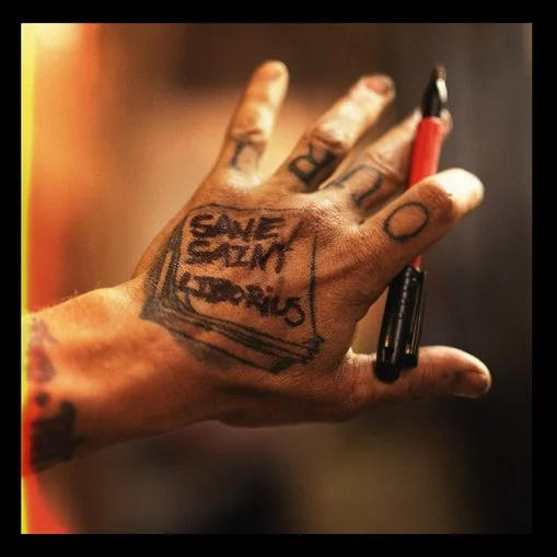 A person's hand with tattoos, holding a red marker, displaying a tattoo that says 'SAVE THE SODIUM' inside a rectangular box. Sk8 Liborius