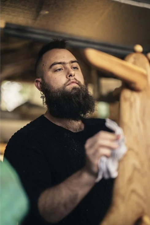 A man with a beard and short hair, wearing a black shirt, polishing or cleaning a wooden sculpture with a cloth, indoors with blurred background.