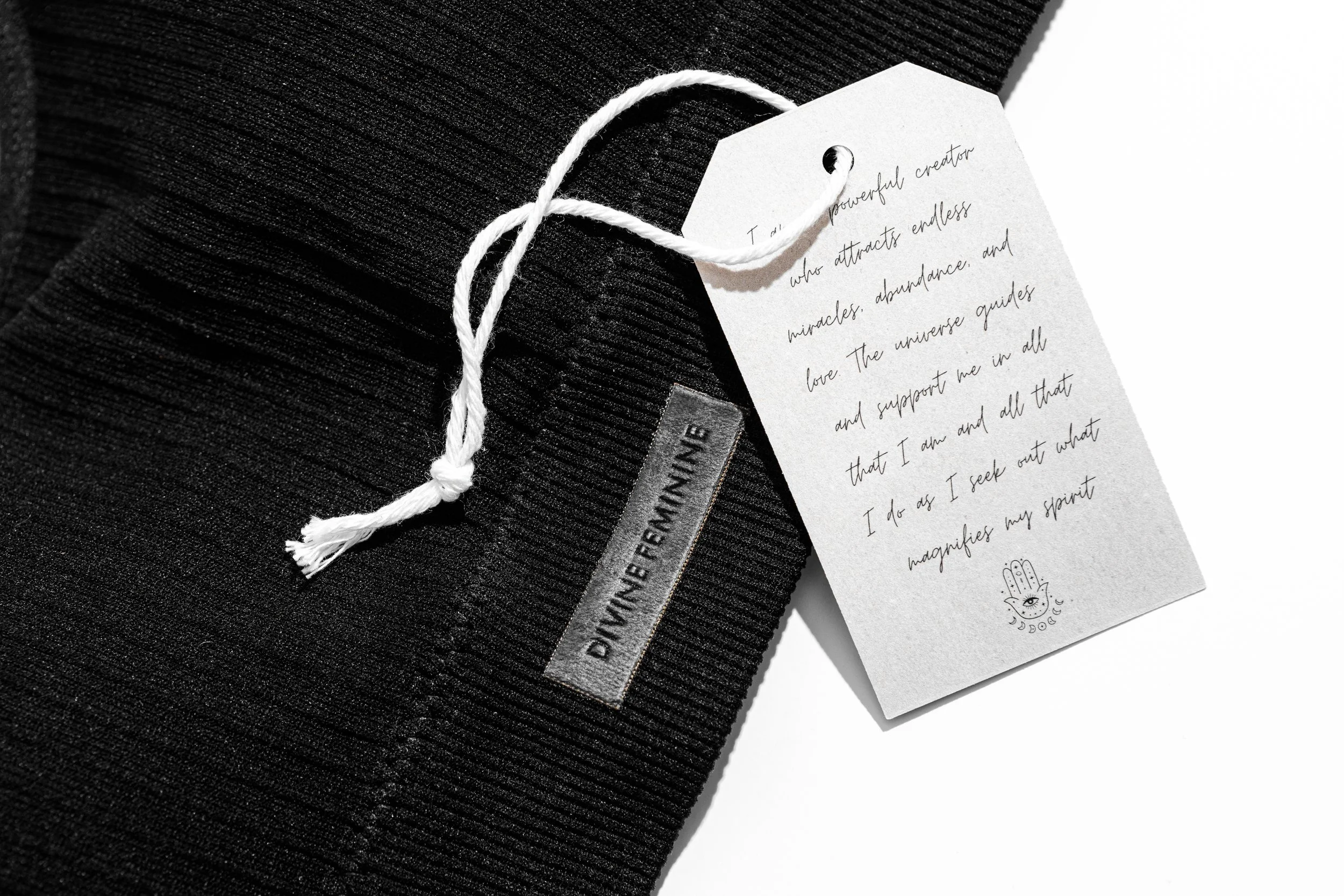 Black textured fabric with a sewn-in label reading "DYNNE FENNINE", a white tag attached with a white string, which has a handwritten note and a small eye and hand illustration at the bottom.