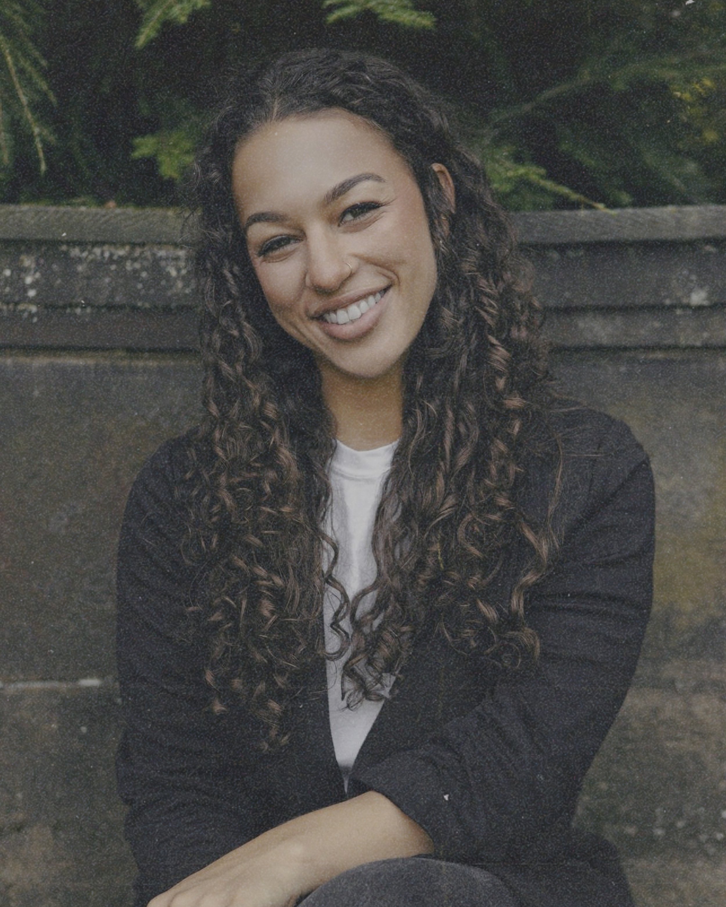 A young woman with long, curly brown hair, smiling, sitting outdoors against a weathered wooden background with greenery.