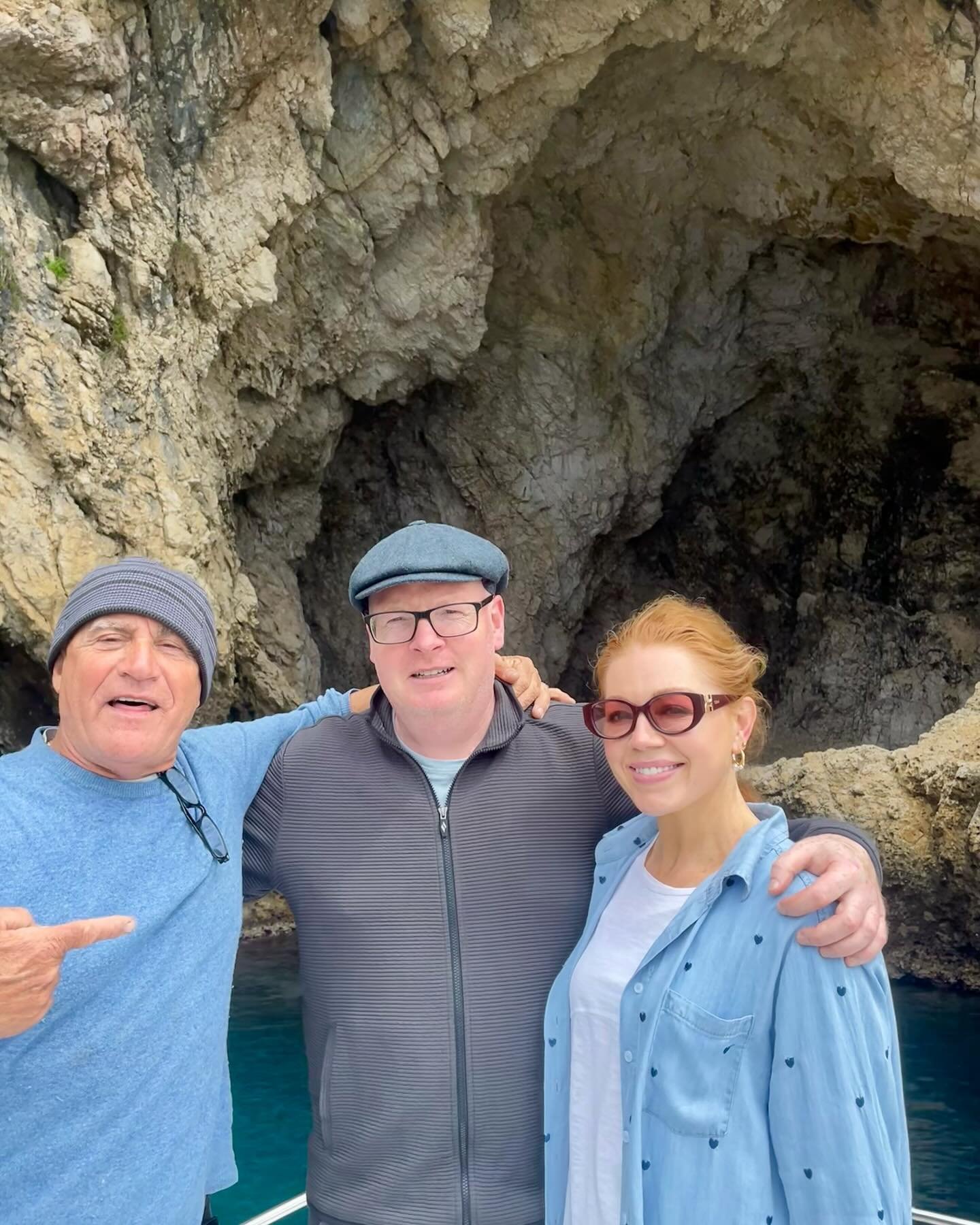 Three people posing in front of a rocky cave entrance by the sea, with one person pointing.