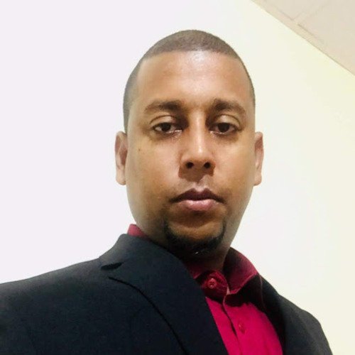 A man with short hair and a serious expression wearing a dark blazer and a maroon shirt, standing against a plain light-colored wall.