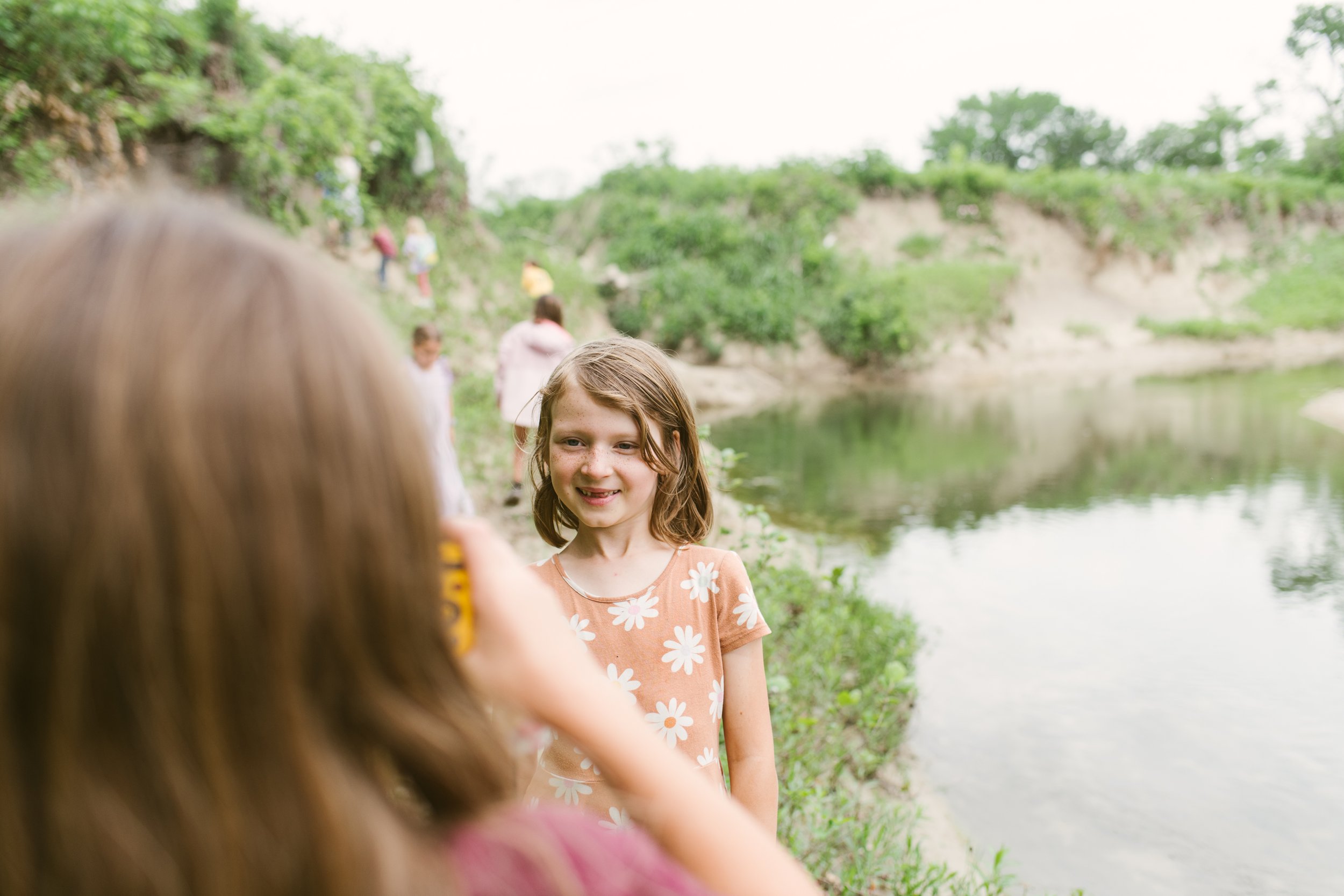 Children by a river, one taking a photo of another, with greenery in the background.