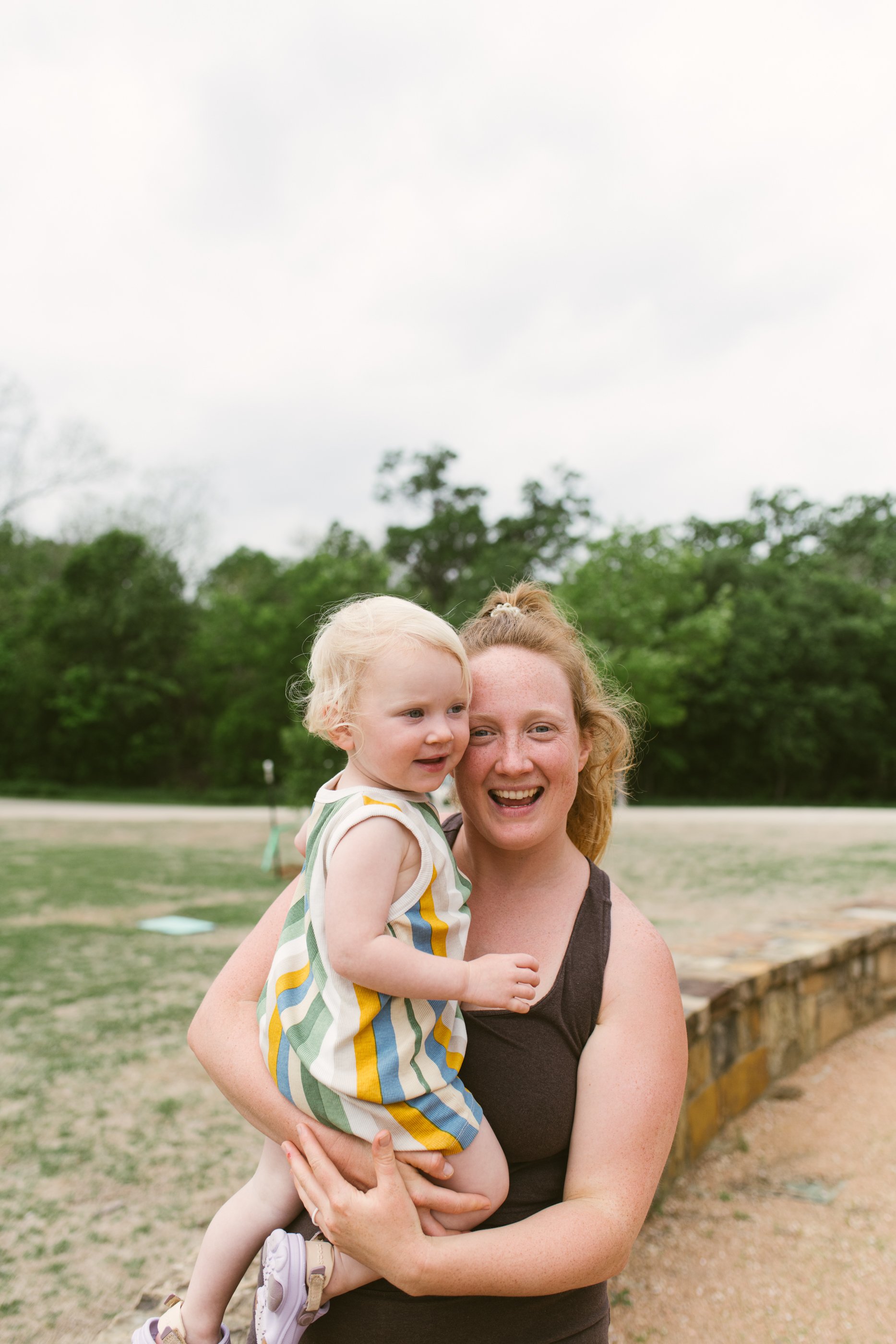 A woman smiling while holding a toddler in a park with trees in the background.