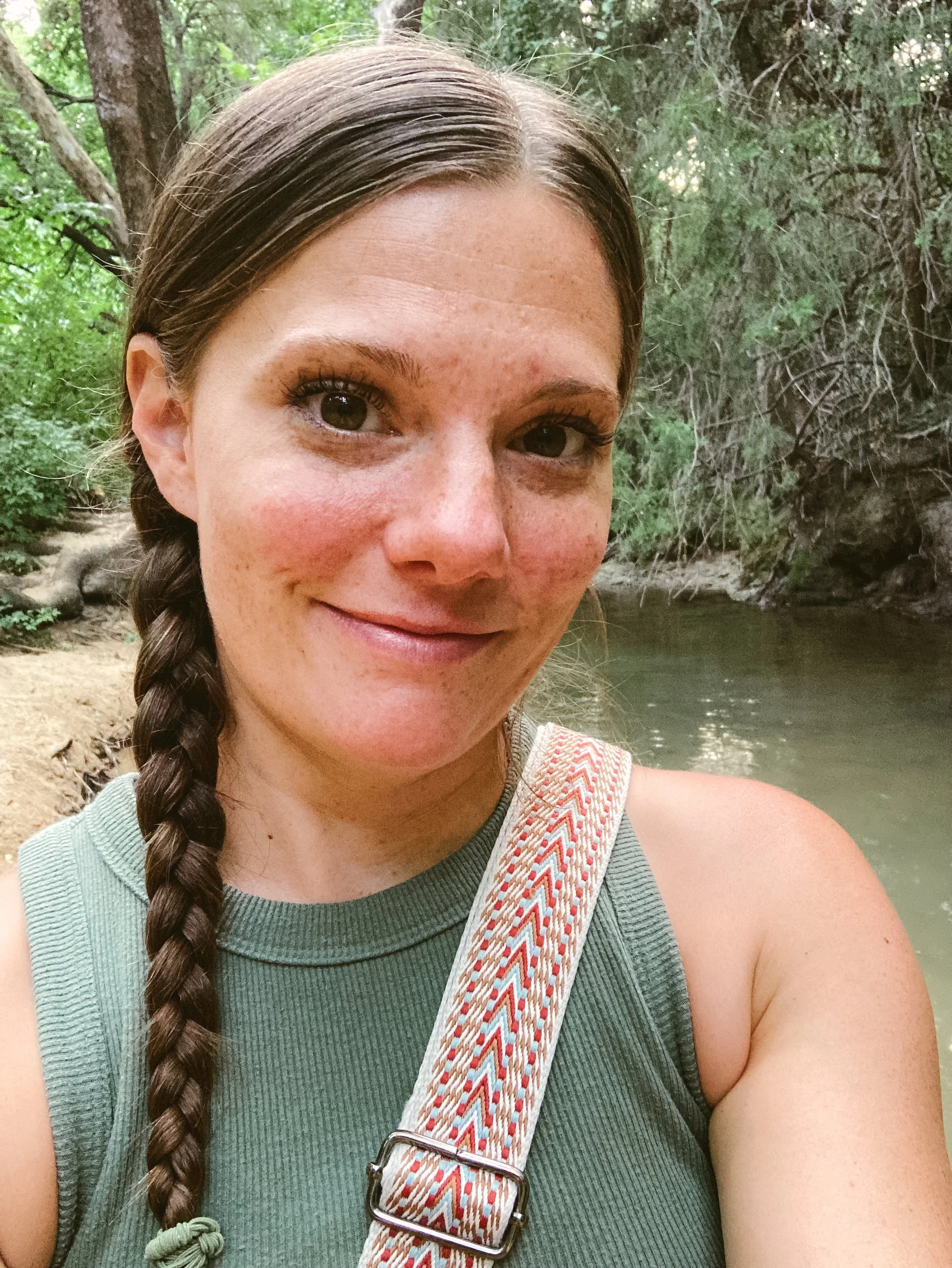 A person with a braided hairstyle, wearing a green sleeveless top and carrying a colorful strap, smiles while standing outdoors near a body of water surrounded by trees.