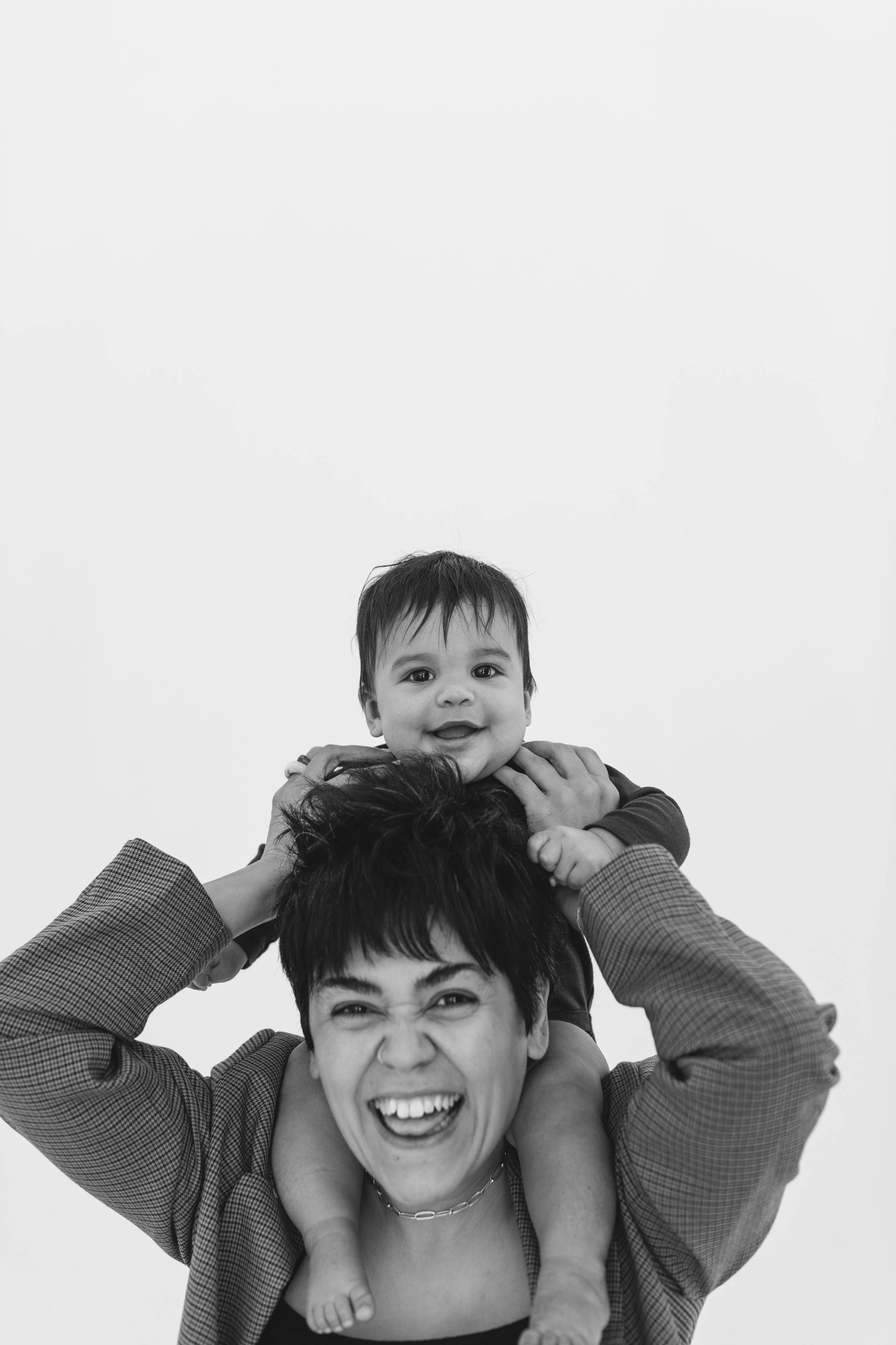 Smiling adult carrying a baby on their shoulders, both laughing, against a plain background.