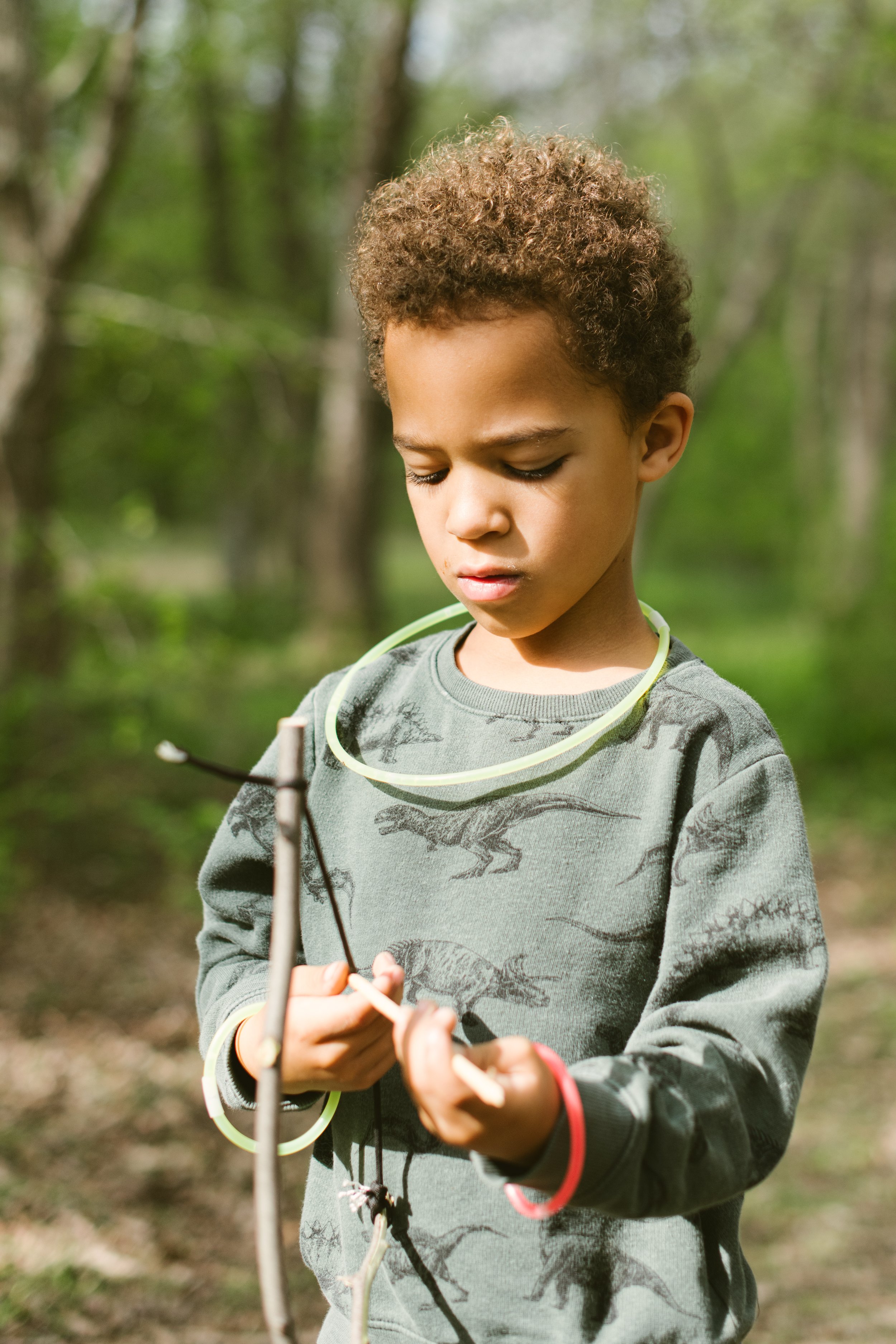 Child in forest wearing dinosaur-print sweater, with glow sticks, holding a stick.