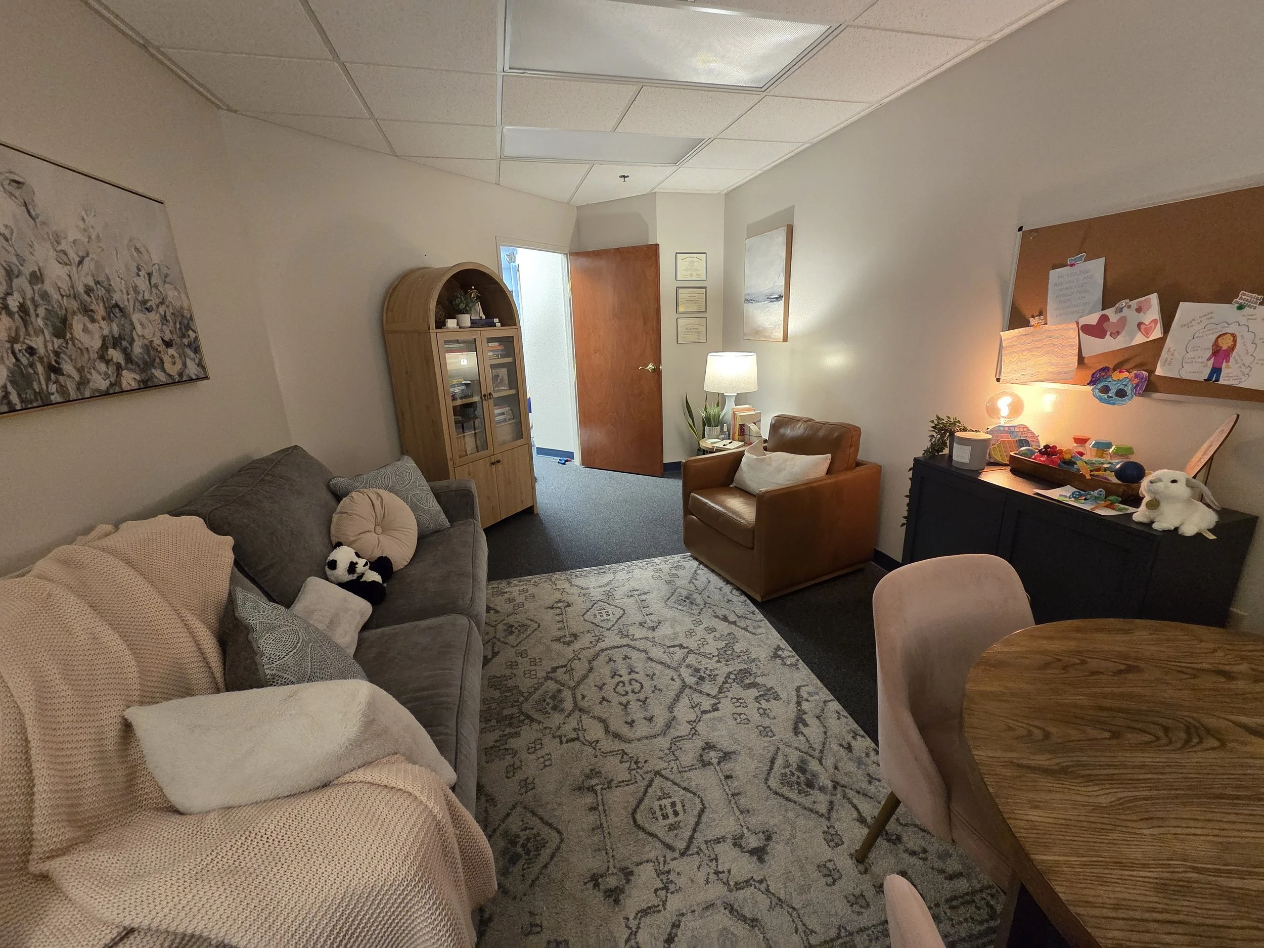 Cozy therapy room with a sofa, armchair, small round table with chairs, decorative rug, lamps, and a mirror. Walls are adorned with artwork and a window allows natural light.