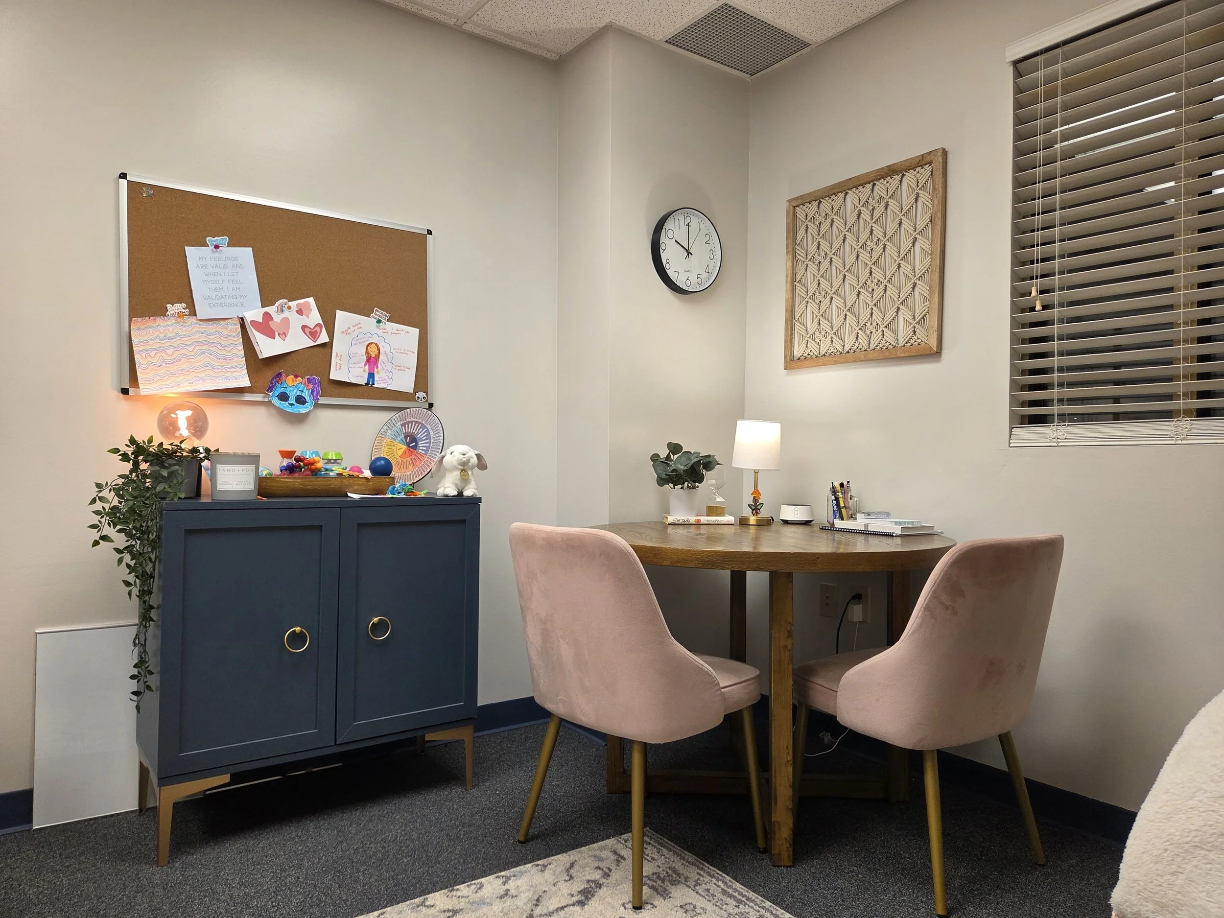Cozy therapy office interior with leather chair, side table, and round dining table with pink chairs. Wall art and indoor plants add decor. Warm lighting enhances the ambiance.