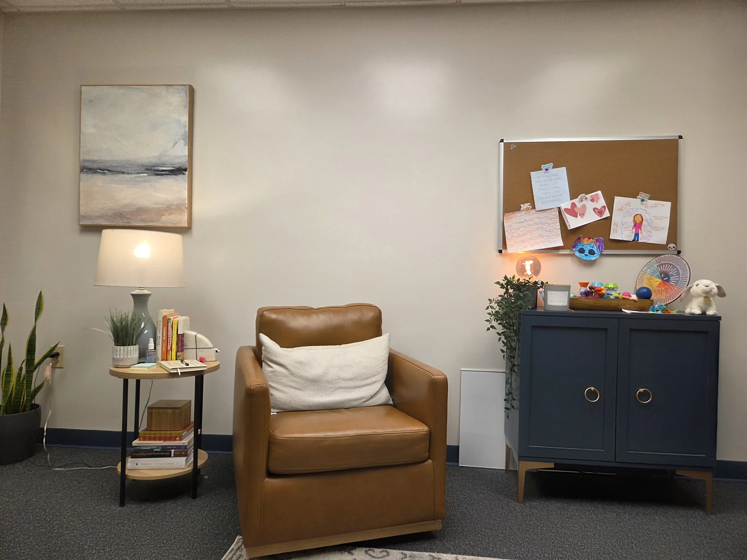 Cozy therapy room with a sofa, armchair, small round table with chairs, decorative rug, lamps, and a mirror. Walls are adorned with artwork and a window allows natural light.