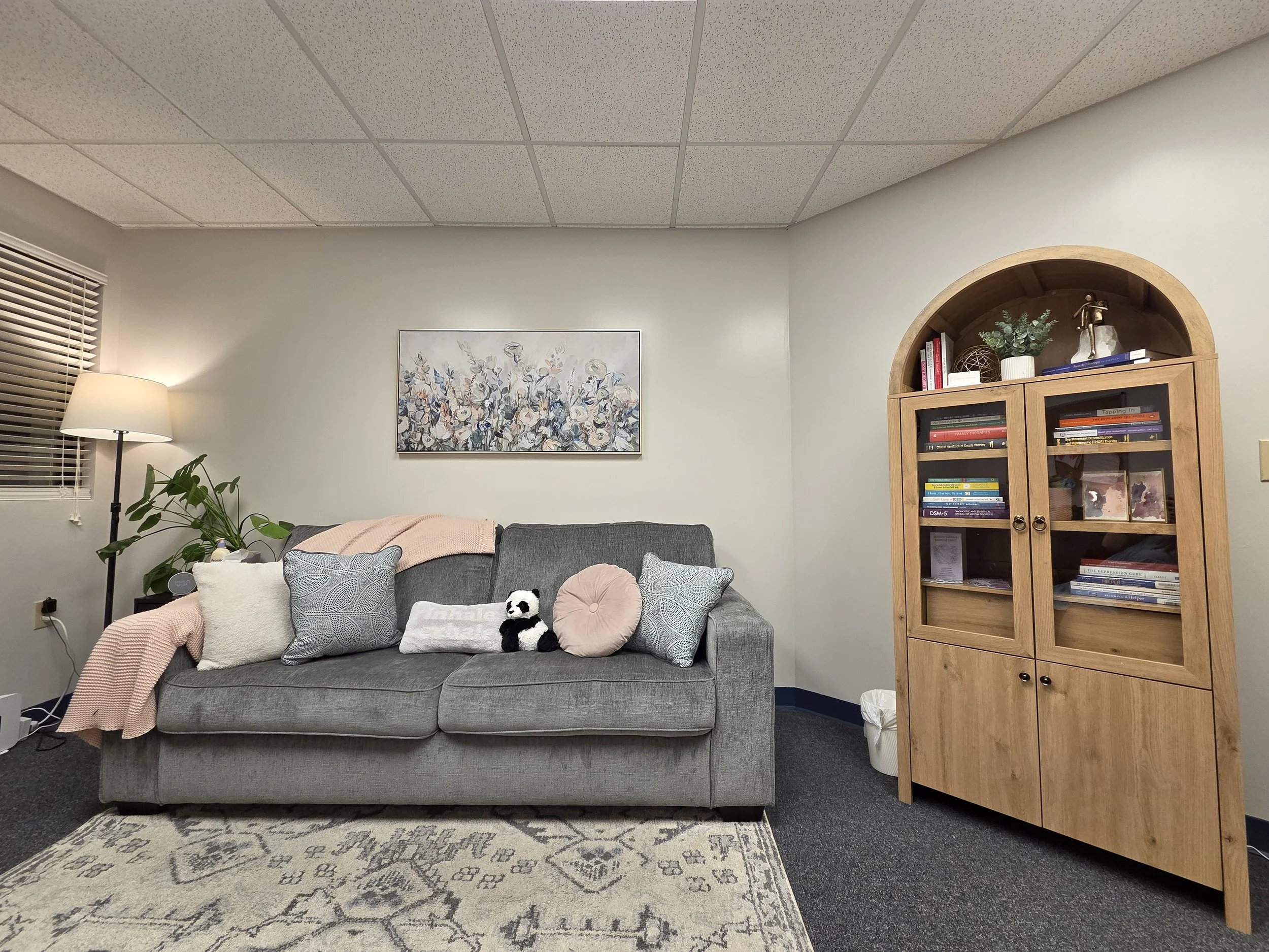 Cozy therapy office with gray sofa, decorative pillows, floral artwork, patterned rug, standing lamp, and cabinet with books and decorative items.