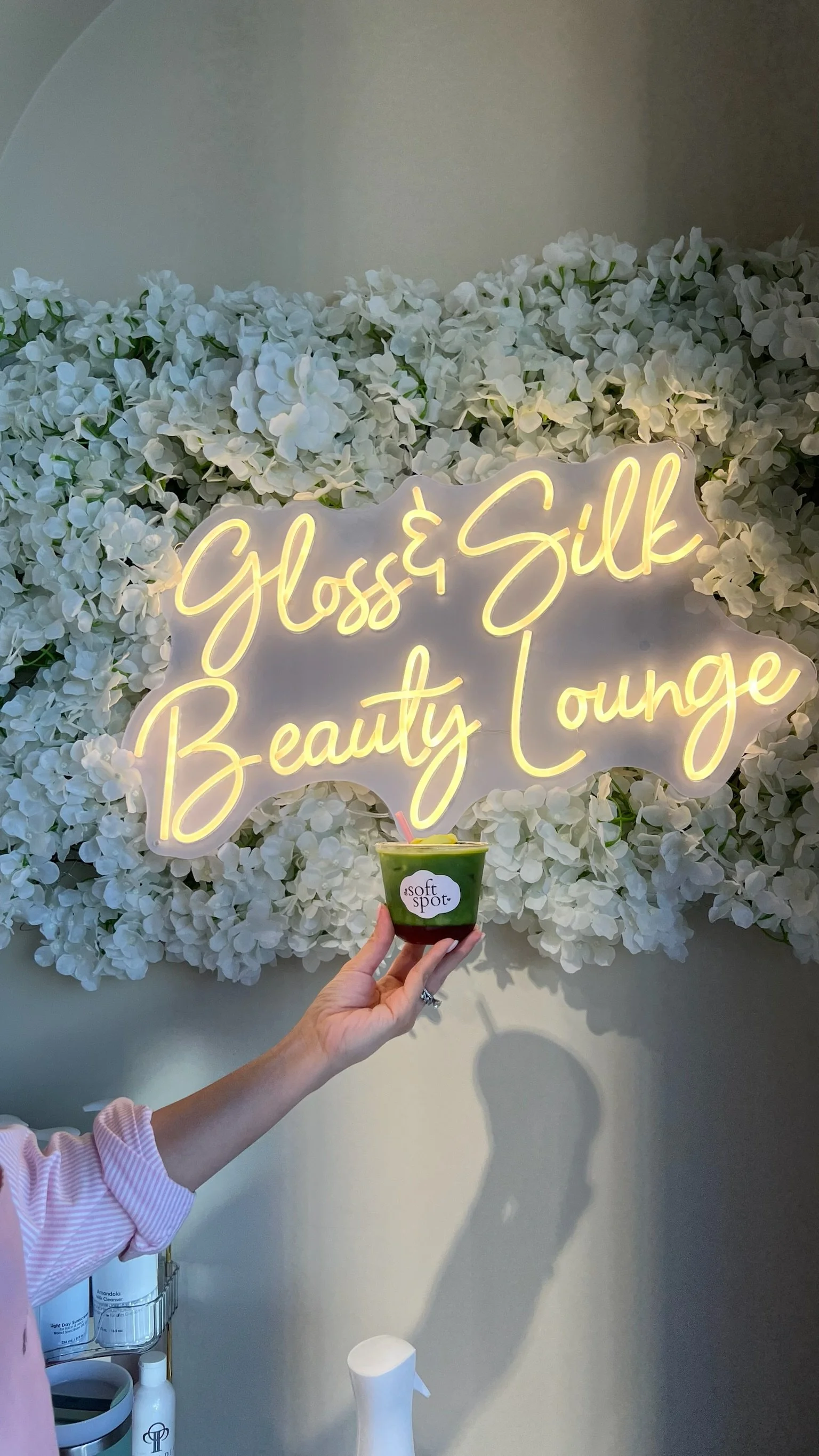 Barista holding a matcha drink prepared by The Soft Spot in front of Gloss&Silk's branded neon light and floral wall sign