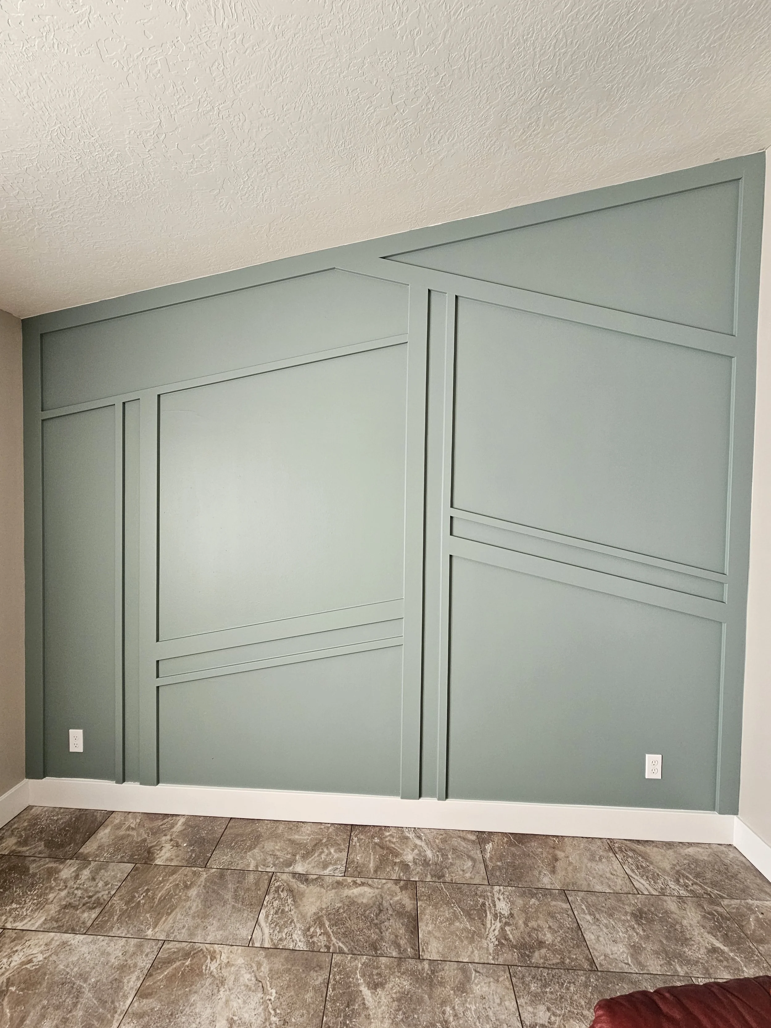 Interior wall painted in light green with rectangular wall panels, located above a beige tile floor and adjacent to a light gray column. Two electrical outlets are visible near the baseboard.