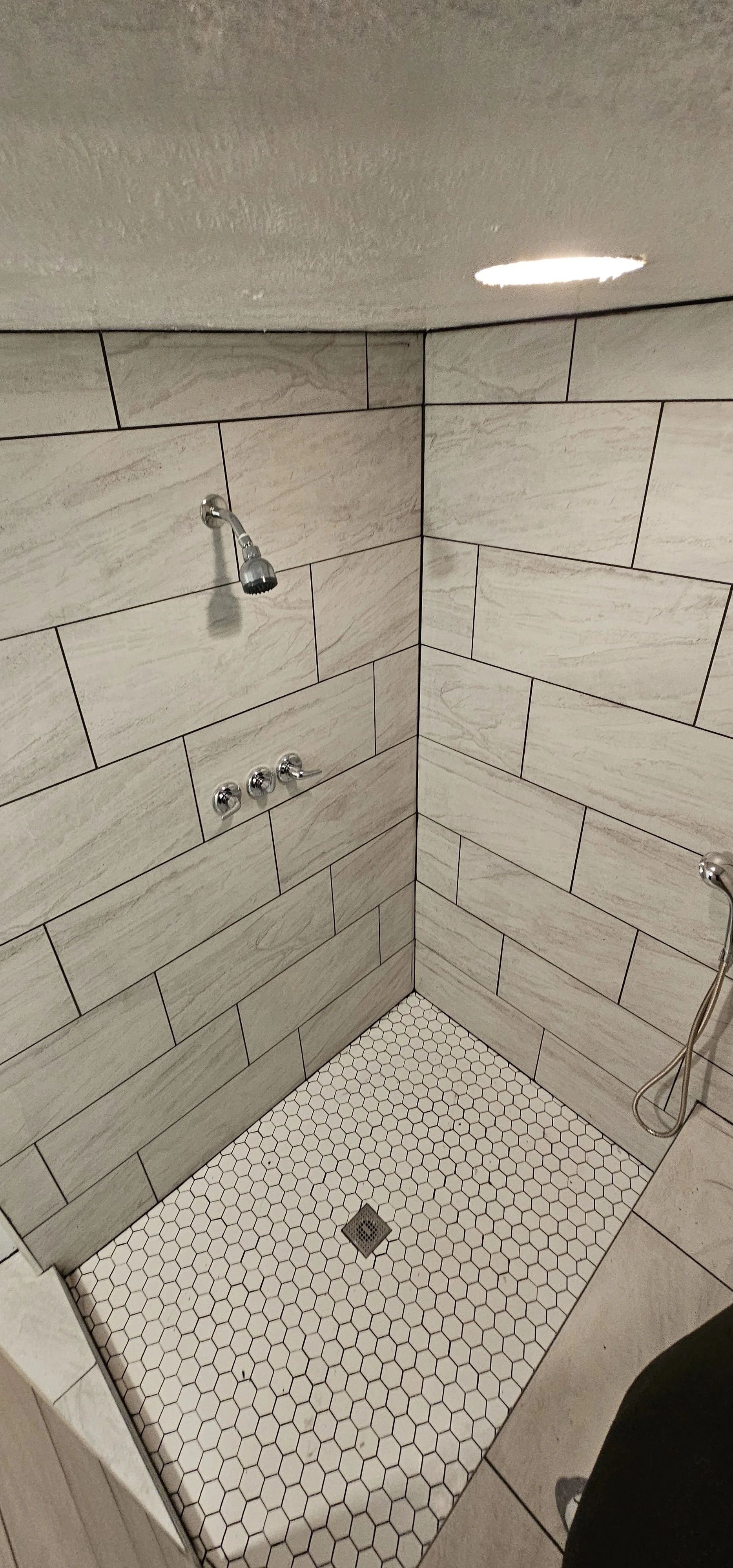 A large walk-in shower tiled with white and gray tiles on the walls, and white hexagonal tiles on the floor.