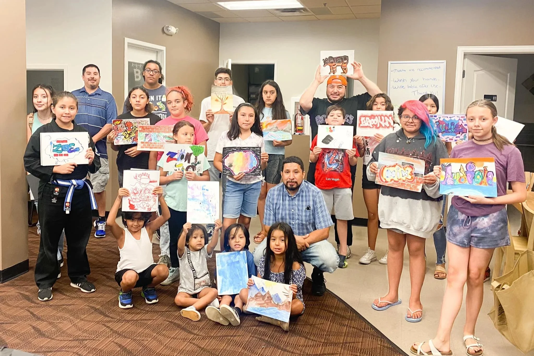 Group of children and adults in a room holding up colorful artwork, with some seated on the floor and others standing behind them. Morrison Road - community.