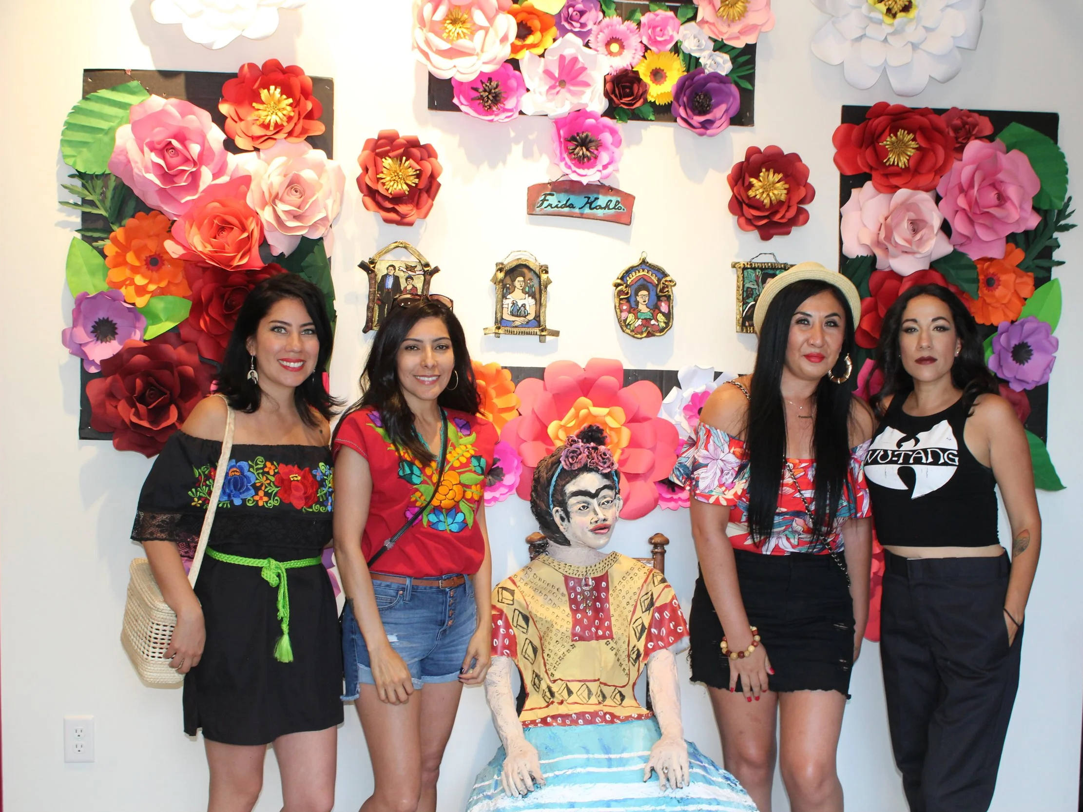 Four women standing in front of colorful paper flowers and Mexican-themed wall art, with a life-sized figure of Frida Kahlo dressed in traditional Mexican attire seated between them.