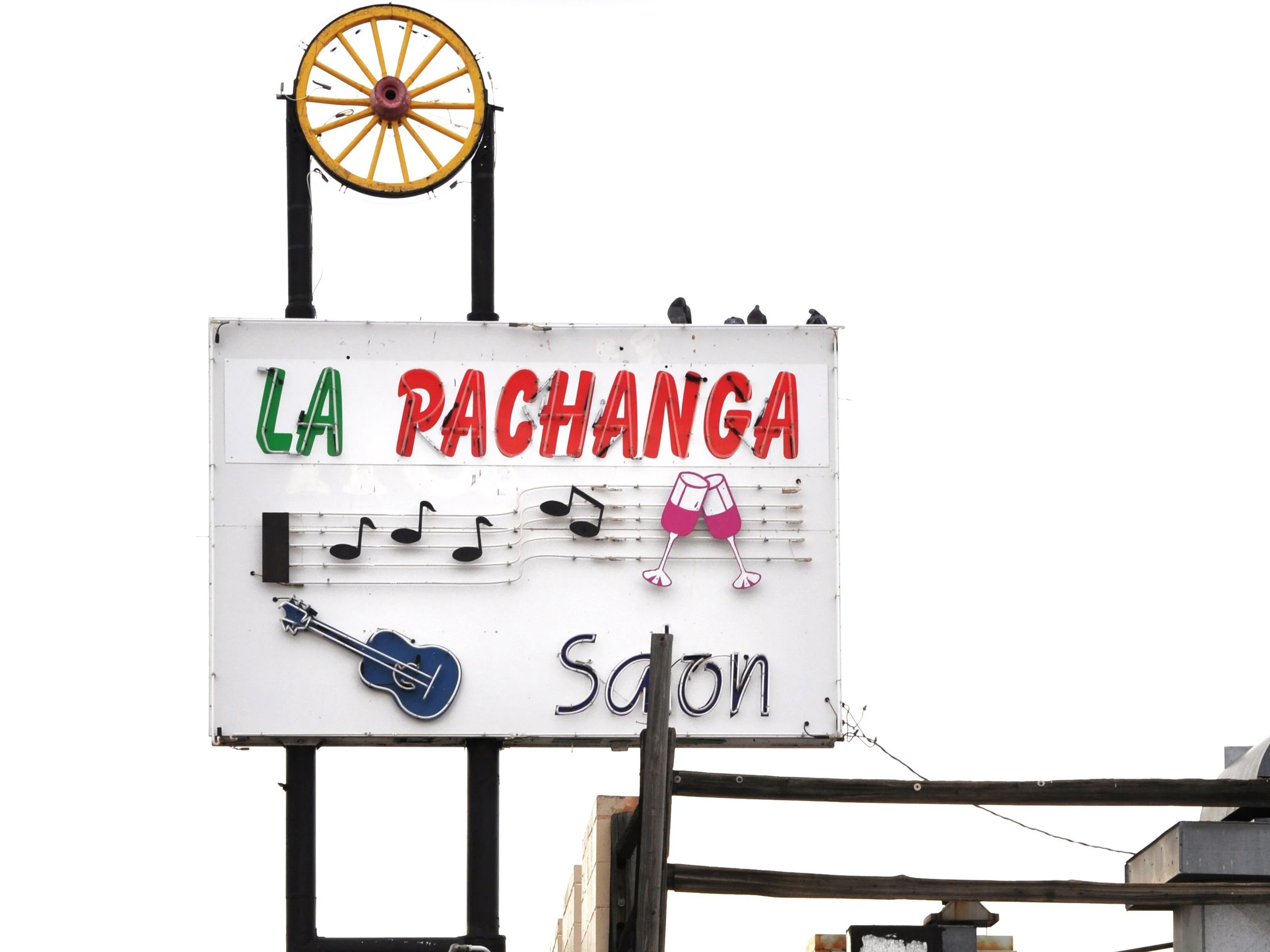 A sign for La Pachanga Salon, featuring musical notes, a guitar, and two clinking wine glasses, with a yellow wagon wheel at the top. The background is white and the sign is mounted on black poles. Morrison Road, Denver Co Business