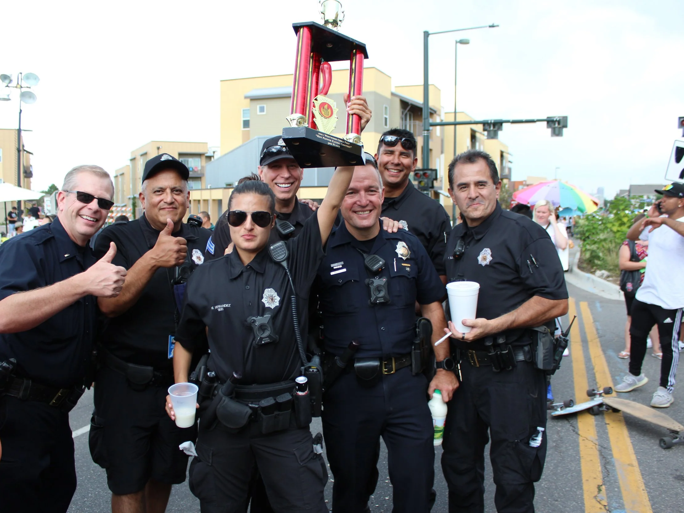 Group of seven police officers celebrating with a trophy at an outdoor event, one officer giving a thumbs-up, others smiling, some holding drinks, a rainbow umbrella and people in the background. Westwood Chile Fest, Denver, CO.