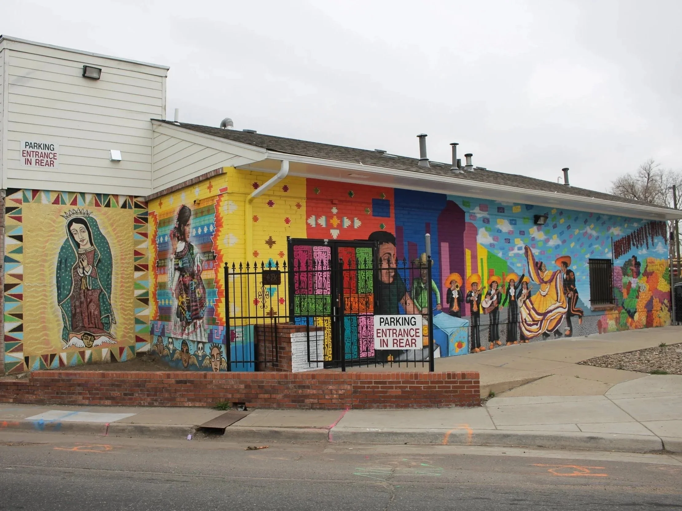 Colorful mural on building wall featuring Mexican cultural scenes, women with sombreros, city skyline, and religious imagery, with two signs reading 'Parking Entrance in Rear' and a black metal gate in front. Morrison Road, Denver Co - mural