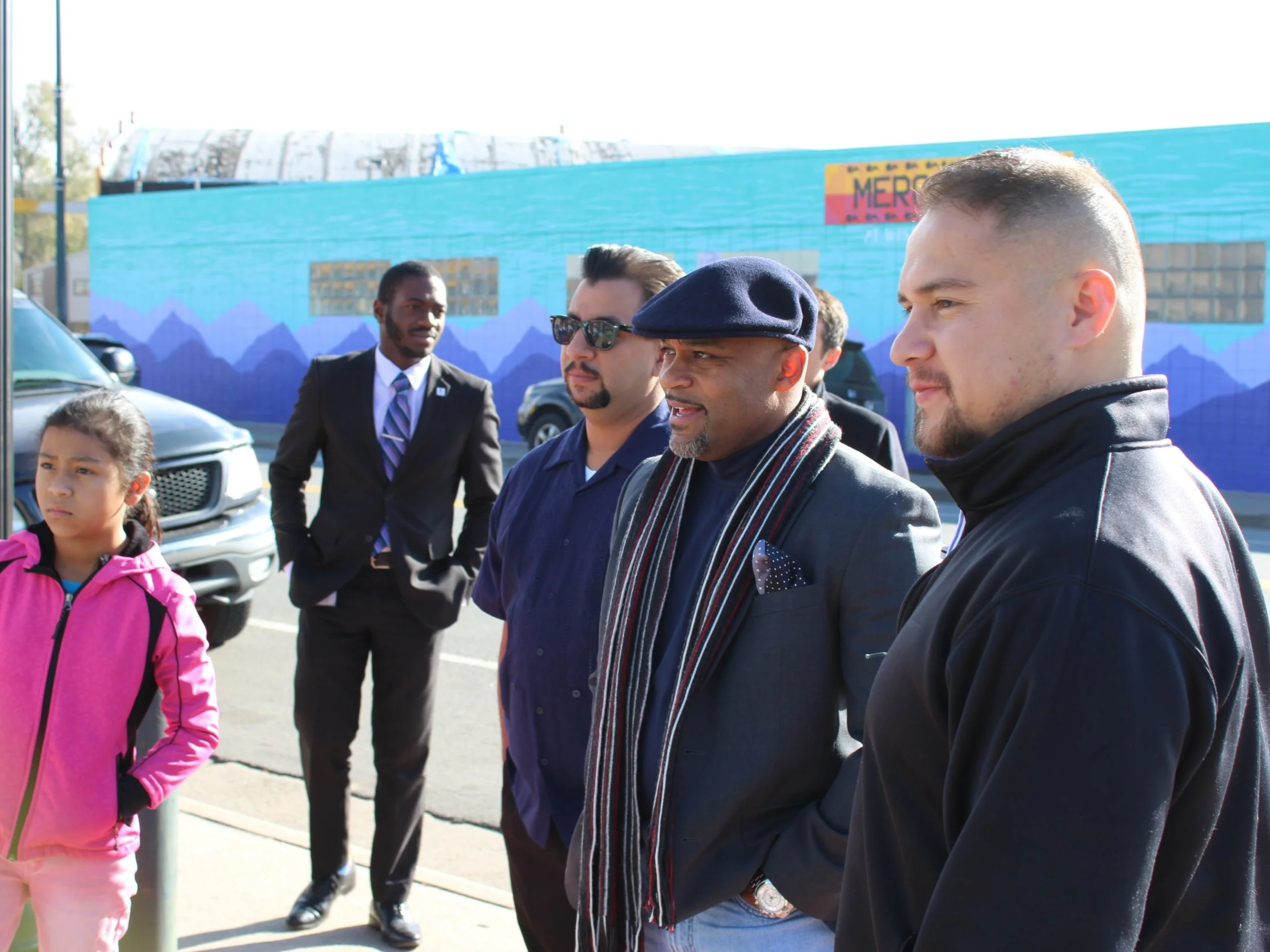 Group of five elected officials, Executive Director Jose Esparza, BuCu West, including a young girl in a pink jacket, standing outdoors in front of a colorful mural with mountains and a bus, with sunlight highlighting their faces.