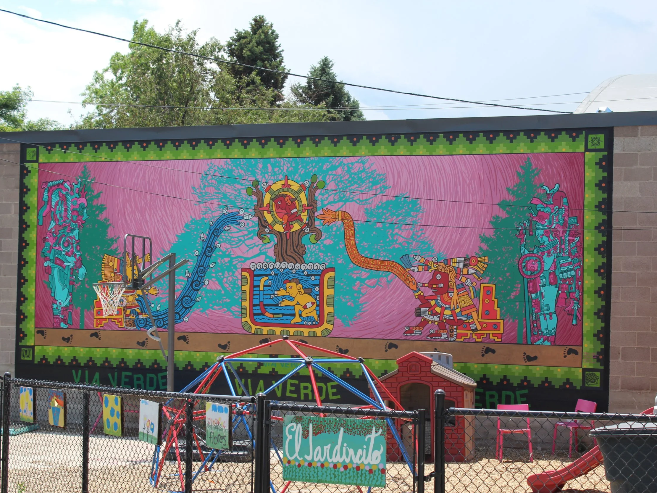 Colorful mural on a playground wall featuring a tree with various abstract and symbolic elements, with a basketball hoop in front and playground equipment below. Morrison Road, Denver, CO Mural