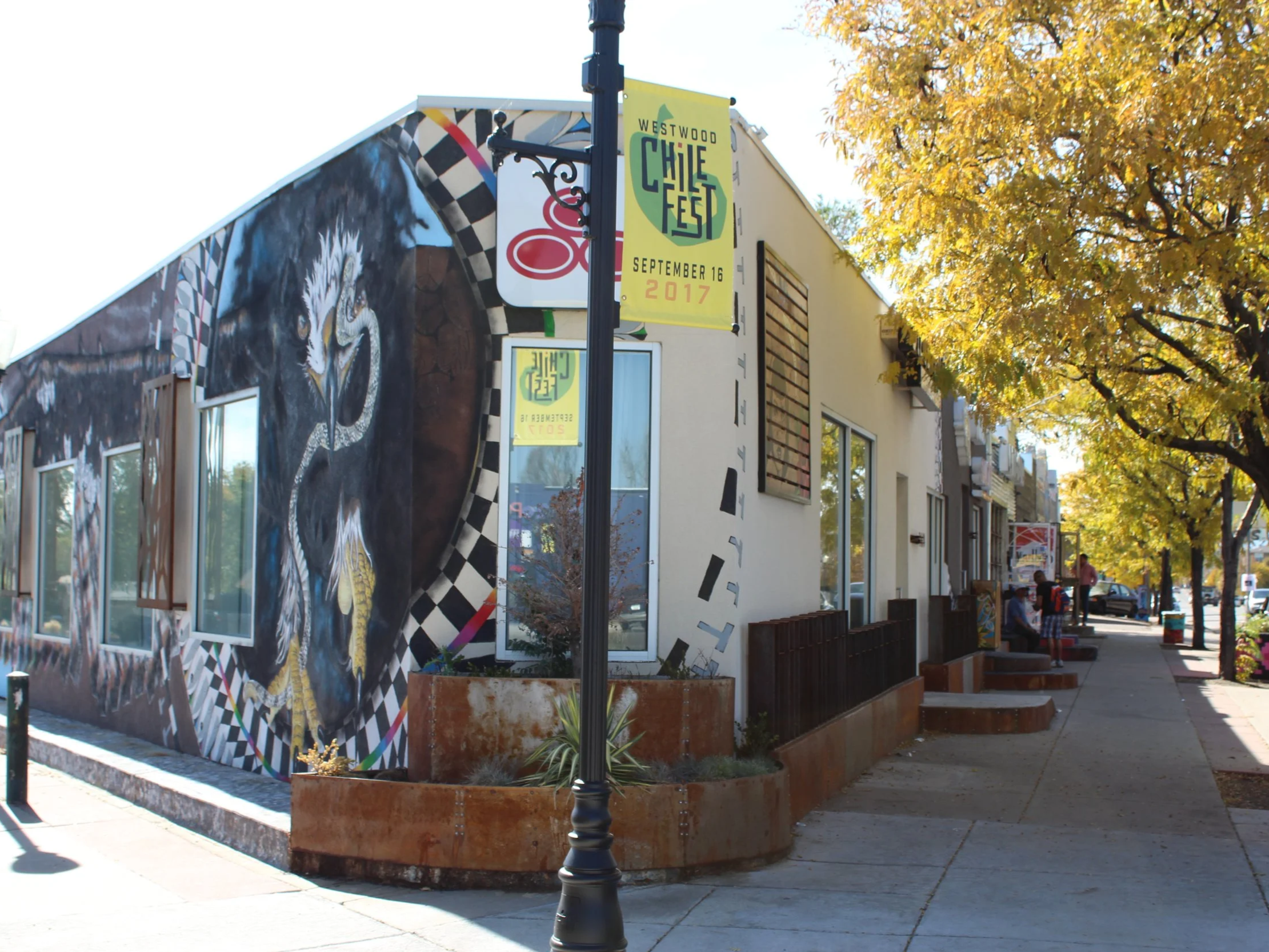 Street view with a mural of a black panther on the side of a building, yellow trees on the sidewalk, and a banner for the Westwood Chile Fest 2017.