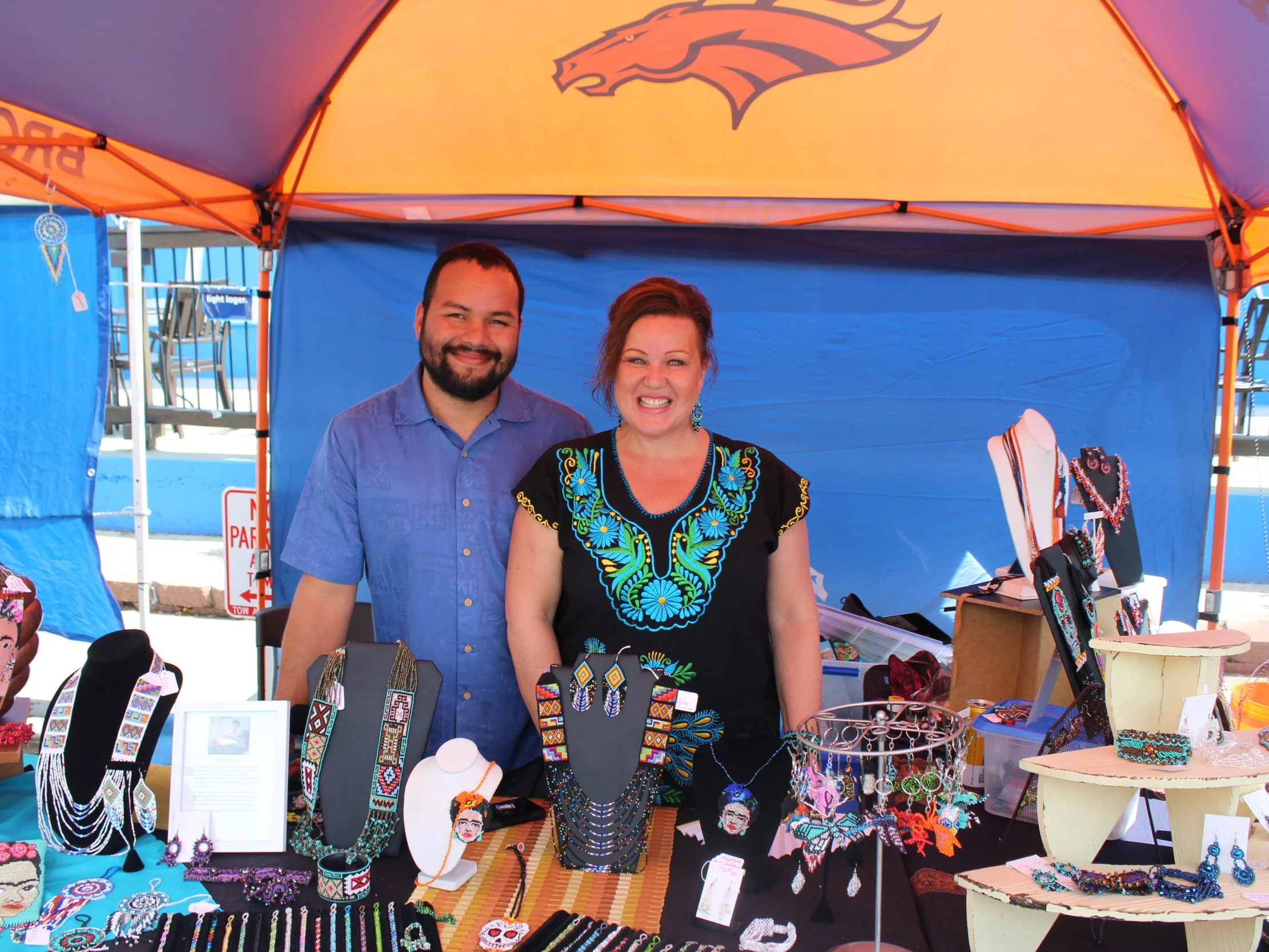 Two people, a man and a woman, standing behind a jewelry display table at an outdoor market stall. They are smiling, and the stall features colorful beaded jewelry, necklaces, earrings, and bracelets. The canopy over the stall has a bright orange and
