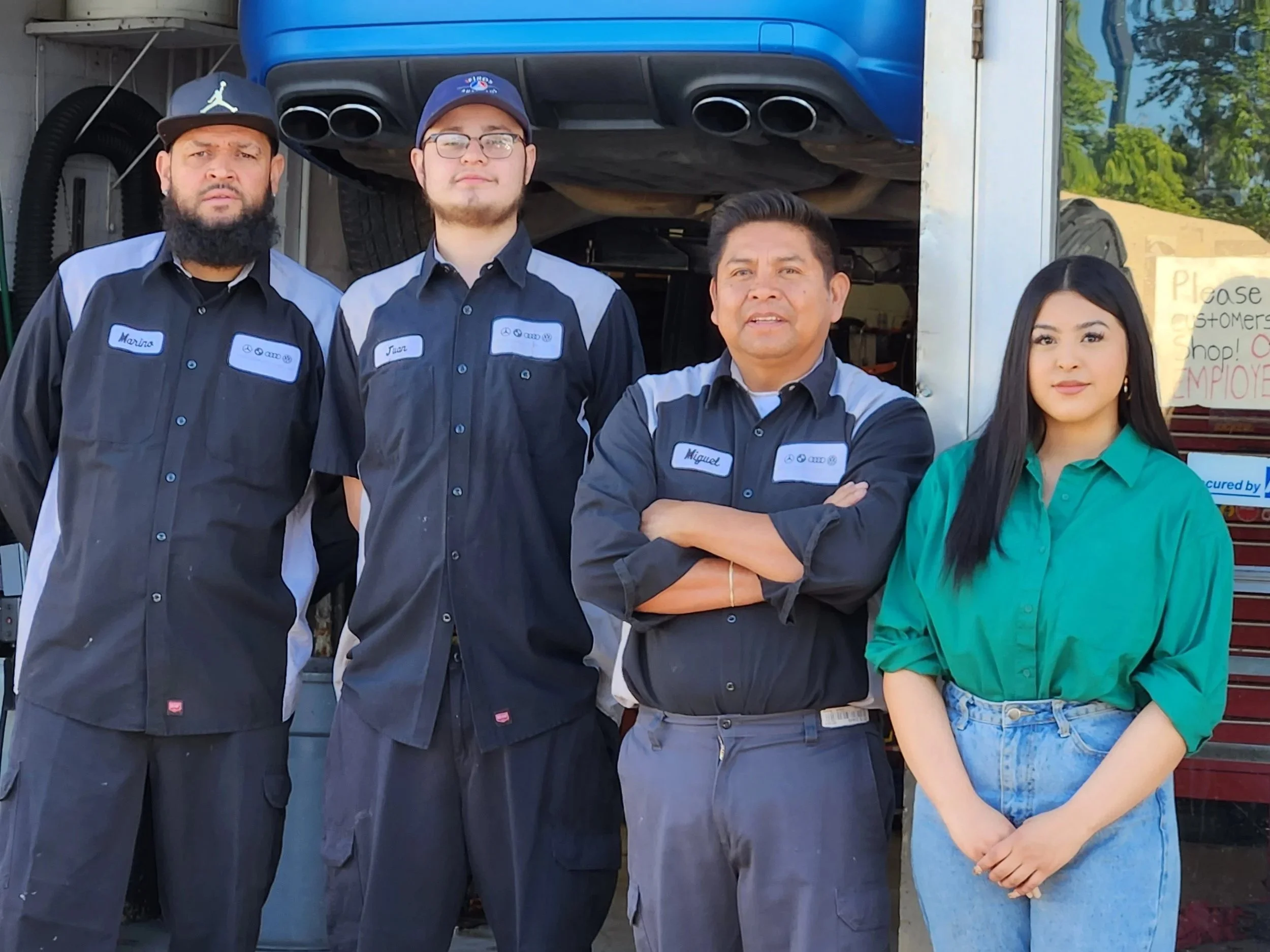 Group of four automotive workers standing in front of a vehicle lift. Three men wearing black and gray uniforms with name tags and a young woman in a green blouse and jeans. Westwood, Denver, CO business owners