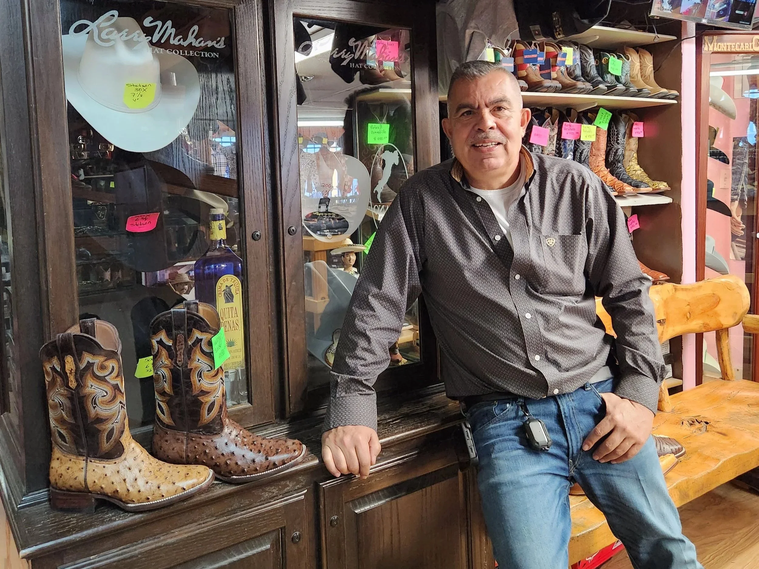 A man in a gray shirt and jeans leaning against a dark wooden display cabinet containing cowboy boots, glass bottles, and other items inside a store. Westwood, Denver, CO Business Owner