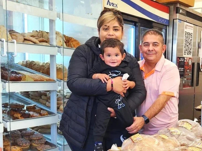 A woman, a young boy, and a man standing in a bakery, with shelves of bread and pastries behind them. The woman is holding the boy, who has a big smile, and the man is smiling next to them. Westwood, Denver, CO business owners.