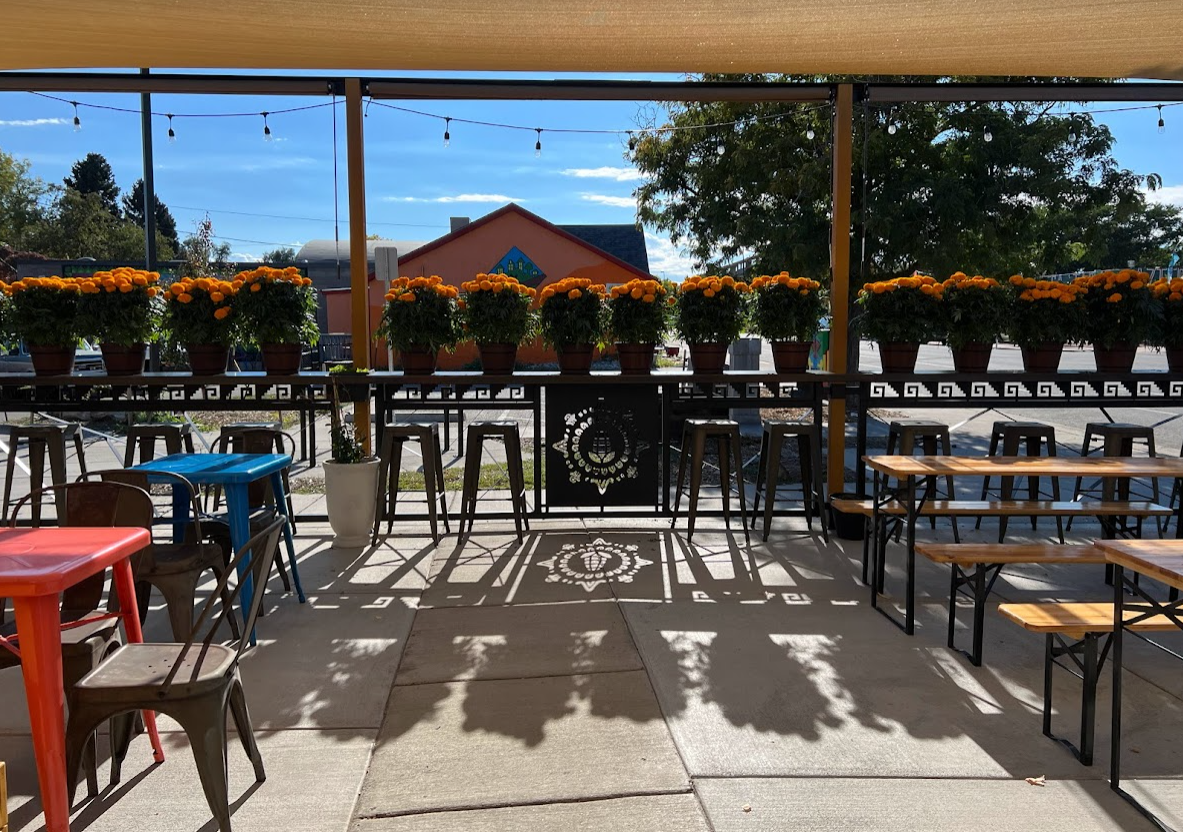 Patio with potted marigold flowers, colorful tables and chairs, and string lights on a sunny day. Cultura Chocolate Cafe- Hechowestwood, Morrison Road, Denver, CO