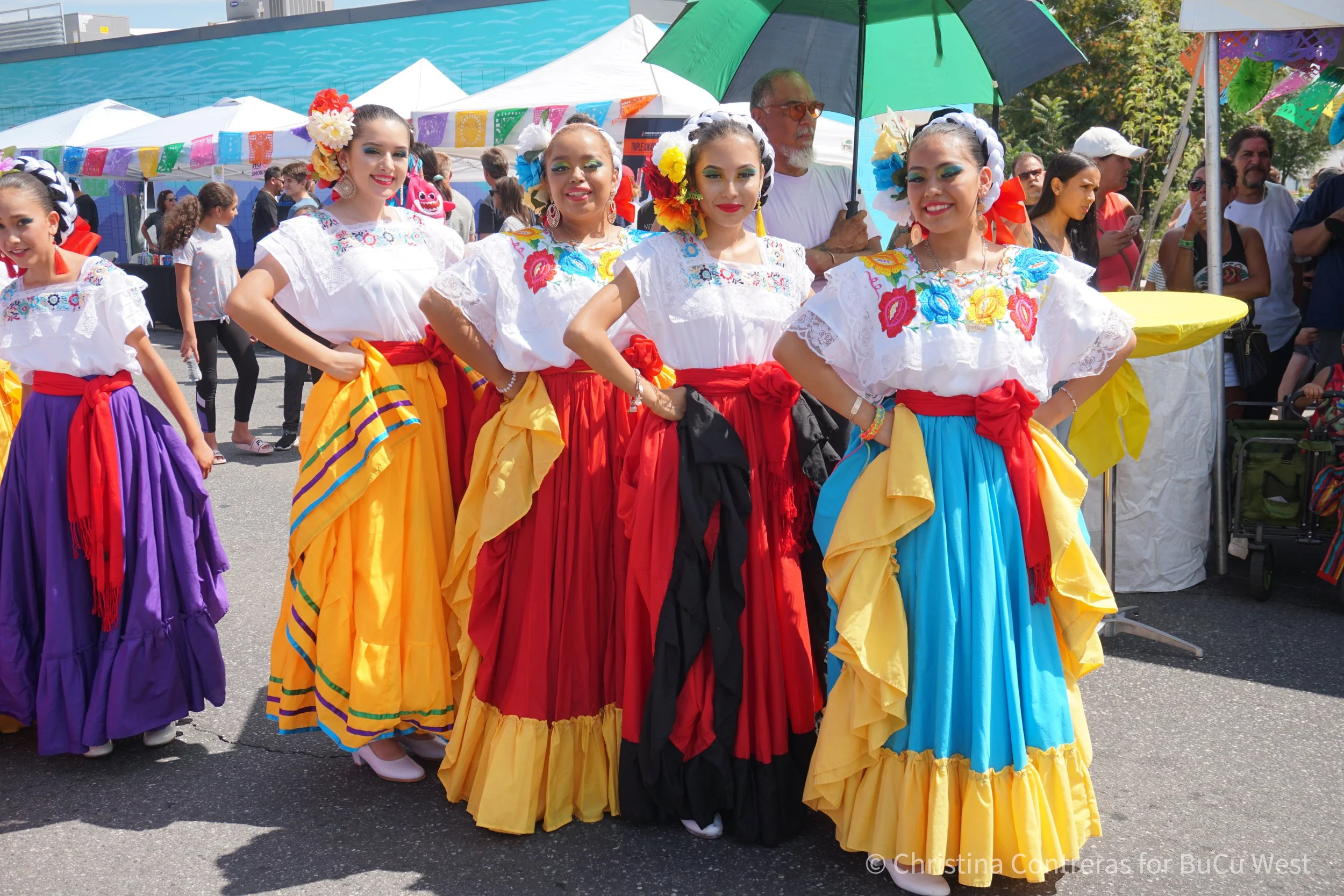 Women dressed in traditional Mexican folkloric dresses with brightly colored skirts and embroidered blouses, standing together at a cultural festival, with tents, other festival attendees, and decorations in the background. Westwood Chile Fest.