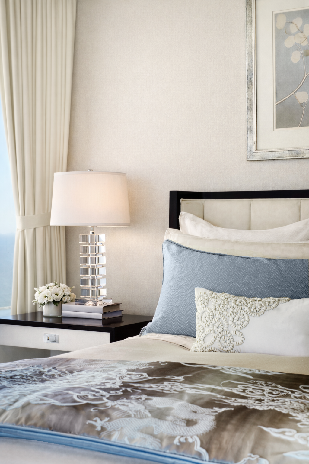 Bedroom with beige curtains, cream-colored wall, framed artwork, black bed frame, white and blue pillows, white bed linens, and a nightstand with a white lamp, a small vase of white flowers, and books.