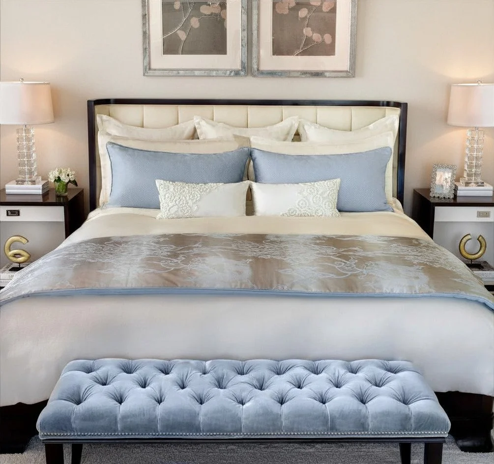 A neatly made bed with white and light blue pillows, a tufted blue bench at the foot, and two matching nightstands with lamps and decorative items.