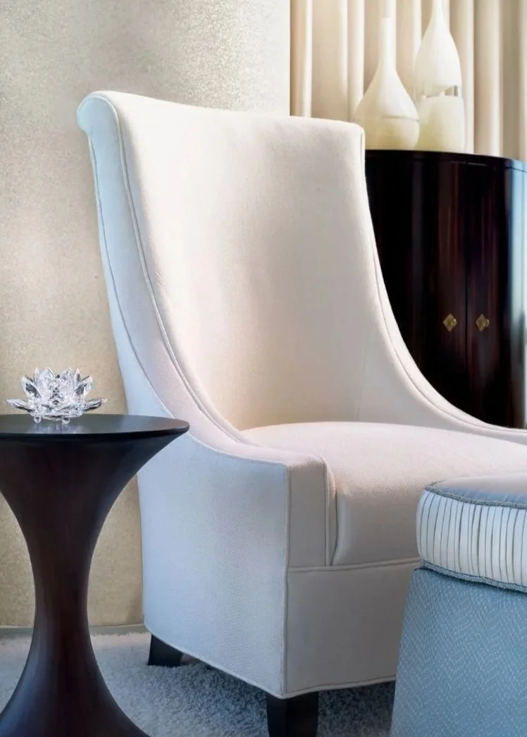 A white high-back armchair next to a black side table with a decorative glass flower on it. In the background, there are cream-colored curtains and white vases on a dark wood cabinet.