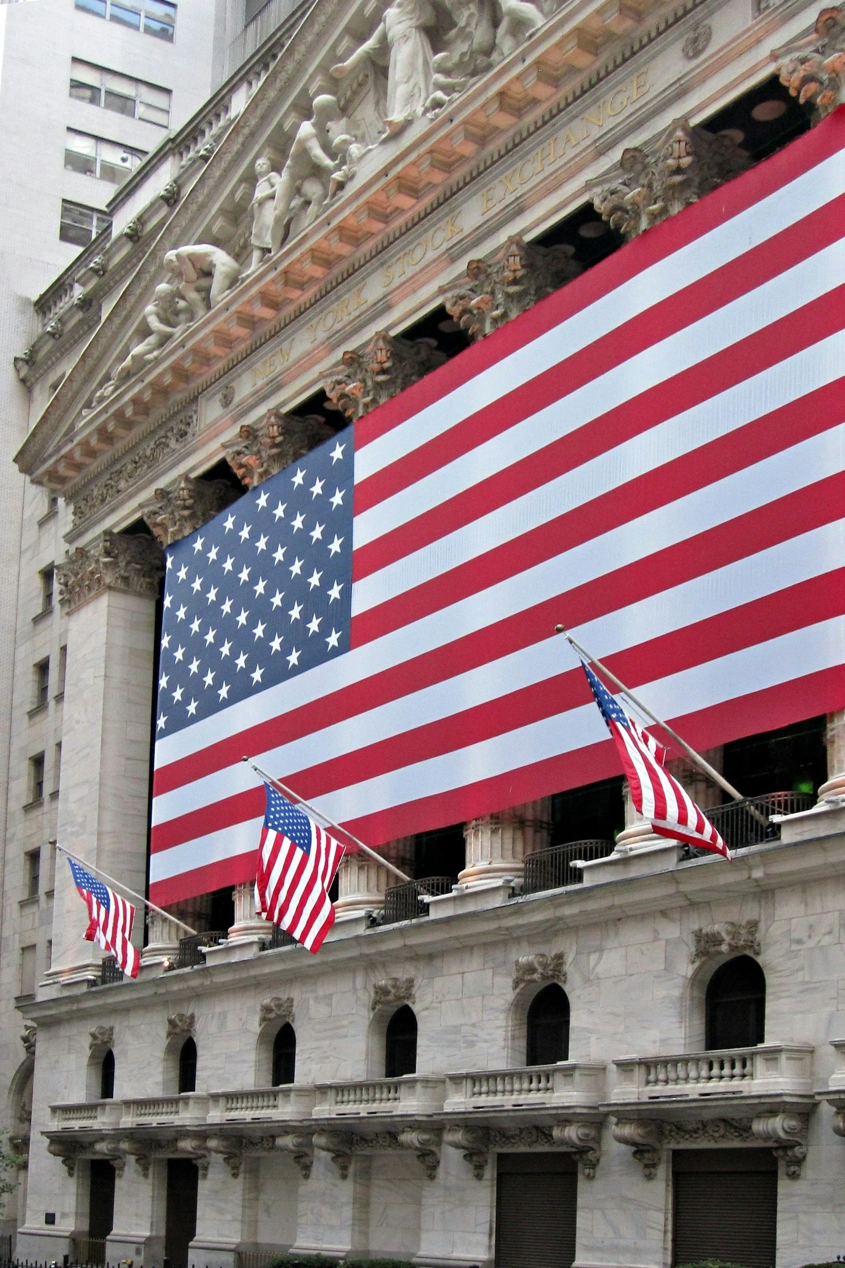 Large American flag hanging from the facade of a classicalStyle building in a city, with smaller flags on either side.