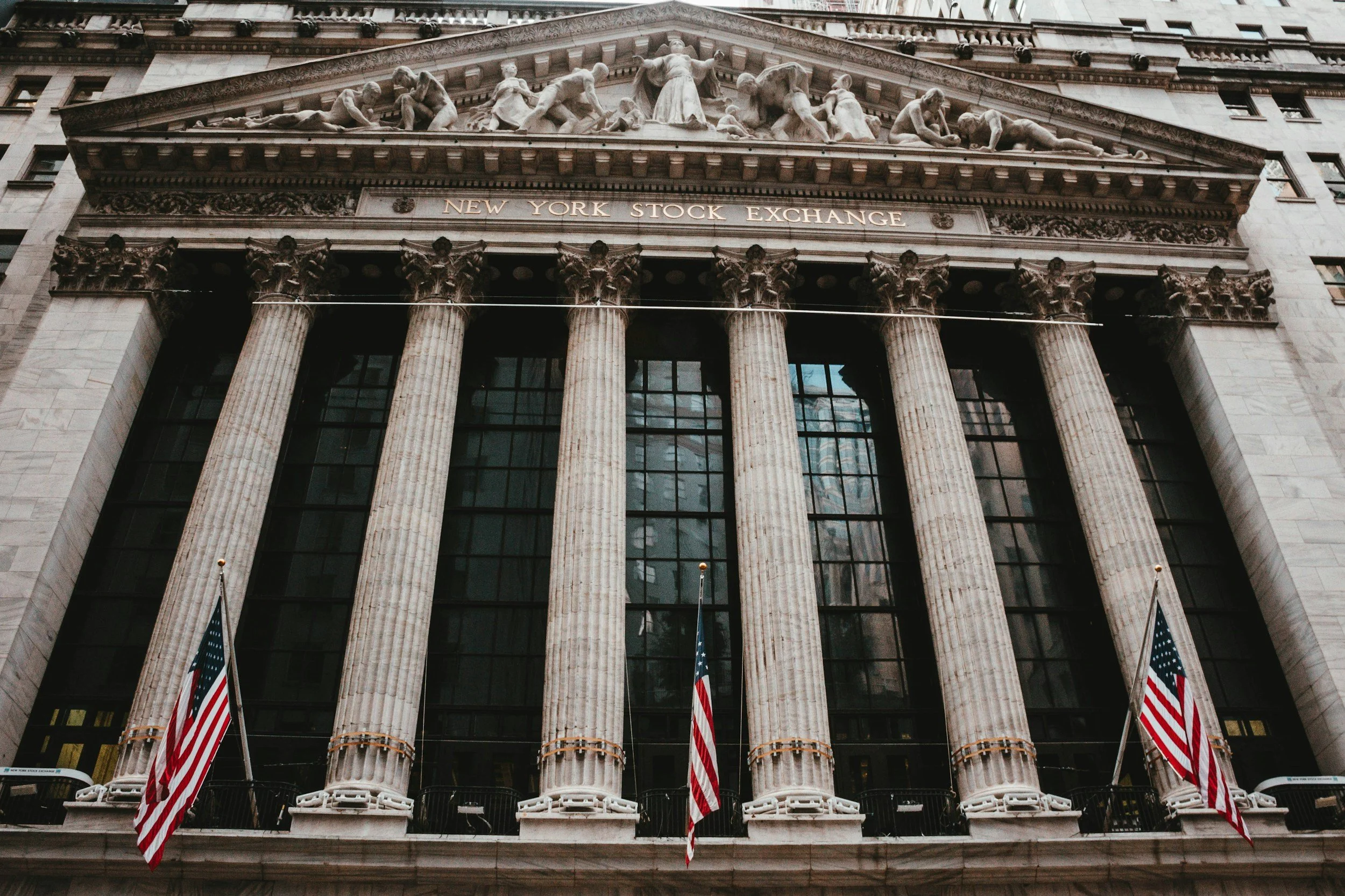 Facade of the New York Stock Exchange building with large columns, flags, and a classical sculpture frieze on top.