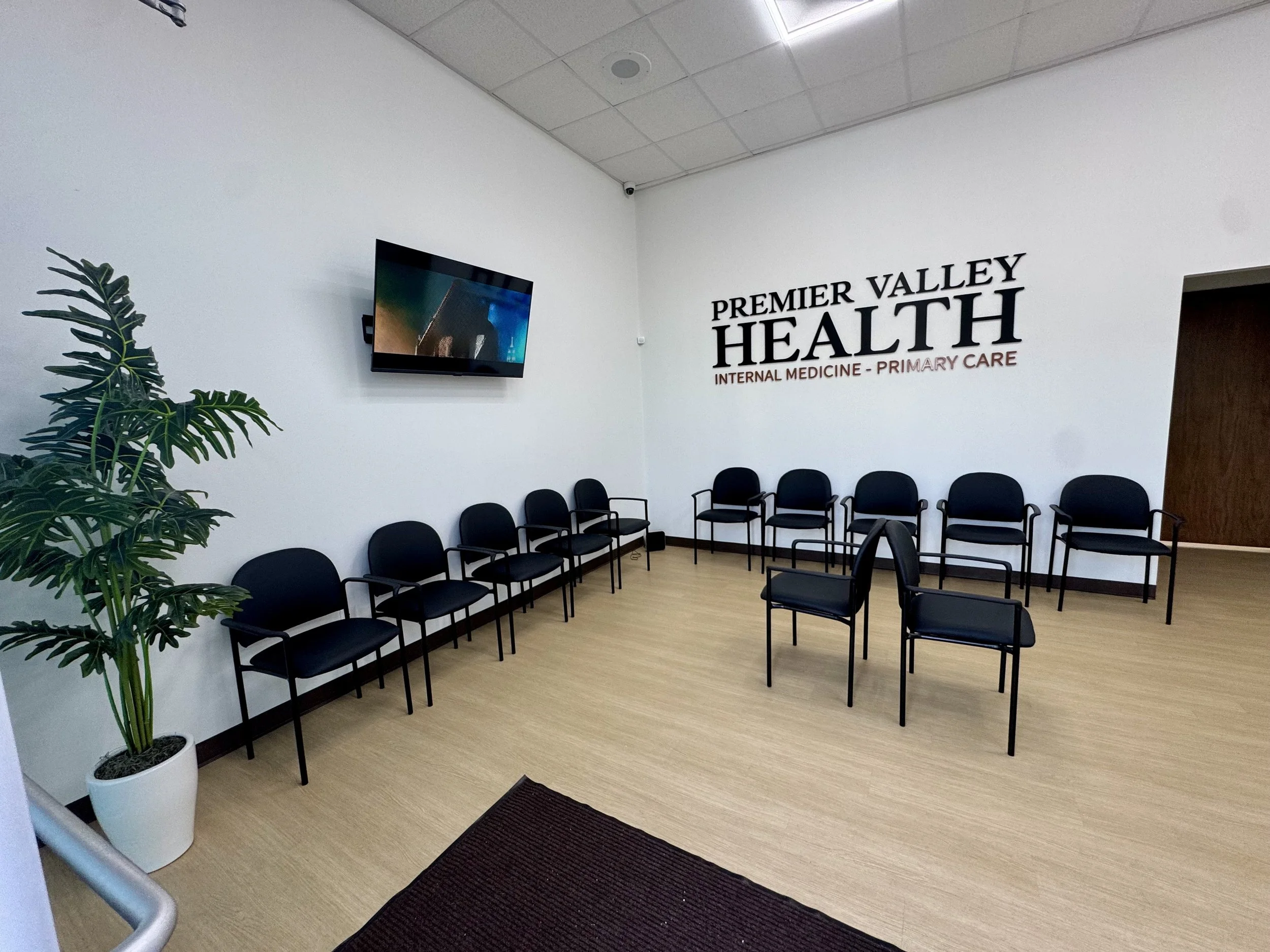 Waiting room with black chairs arranged in two rows, a potted plant in the corner, a wall-mounted television, and a wall with the sign 'Premier Valley Health Internal Medicine - Primary Care' in black and red letters.