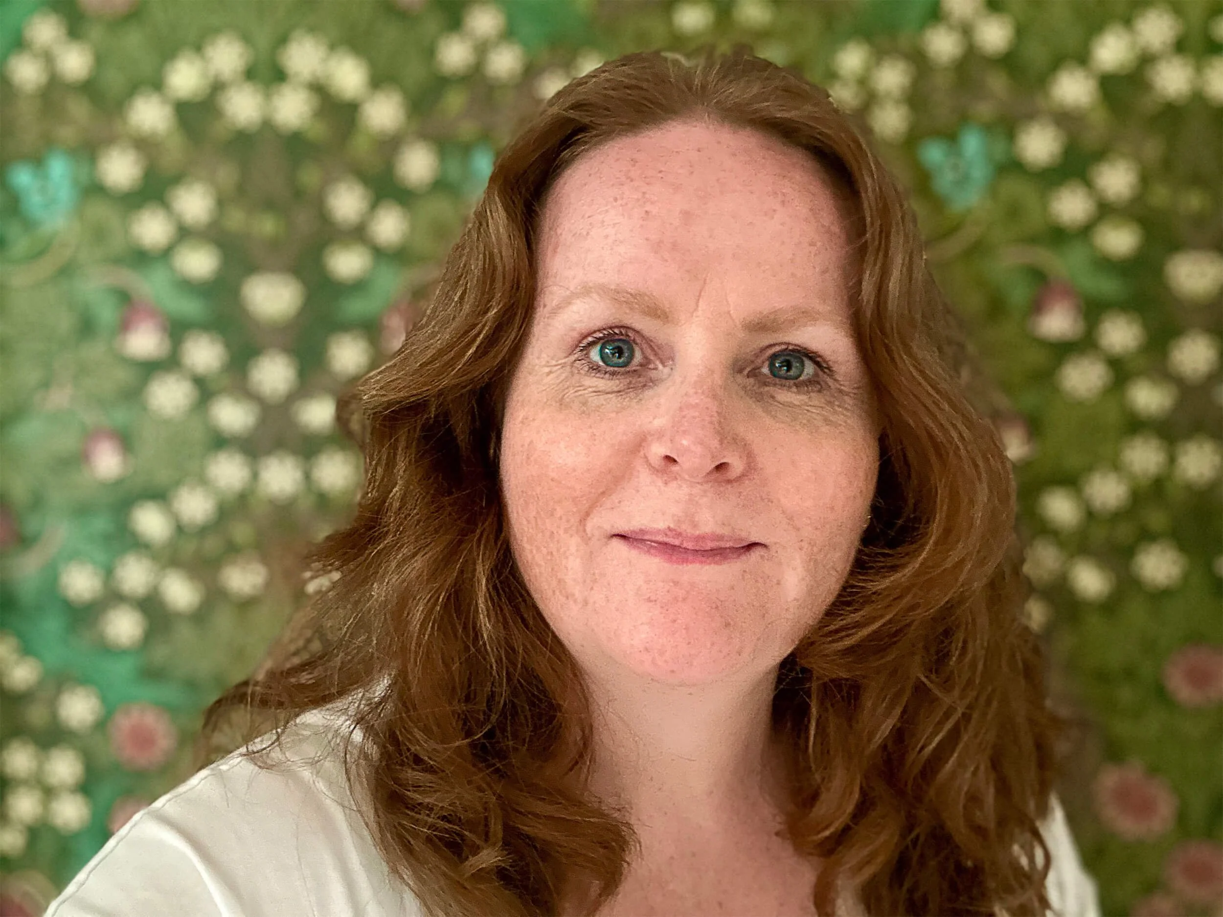 A woman with red hair and blue eyes smiling in front of a floral patterned background.