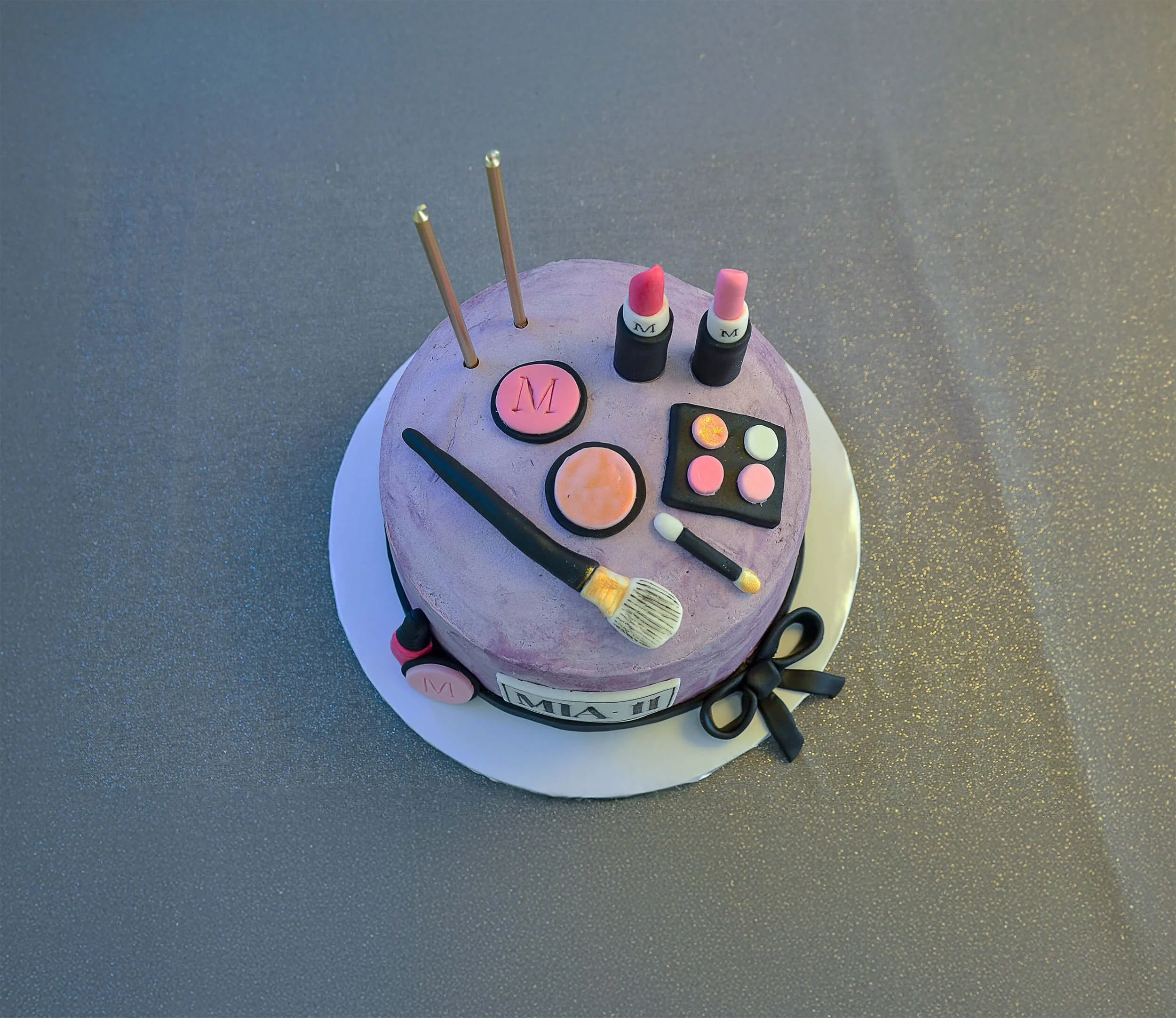 A round makeup-themed cake decorated with lipstick, makeup brushes, eyeshadow palette, and makeup containers on top.