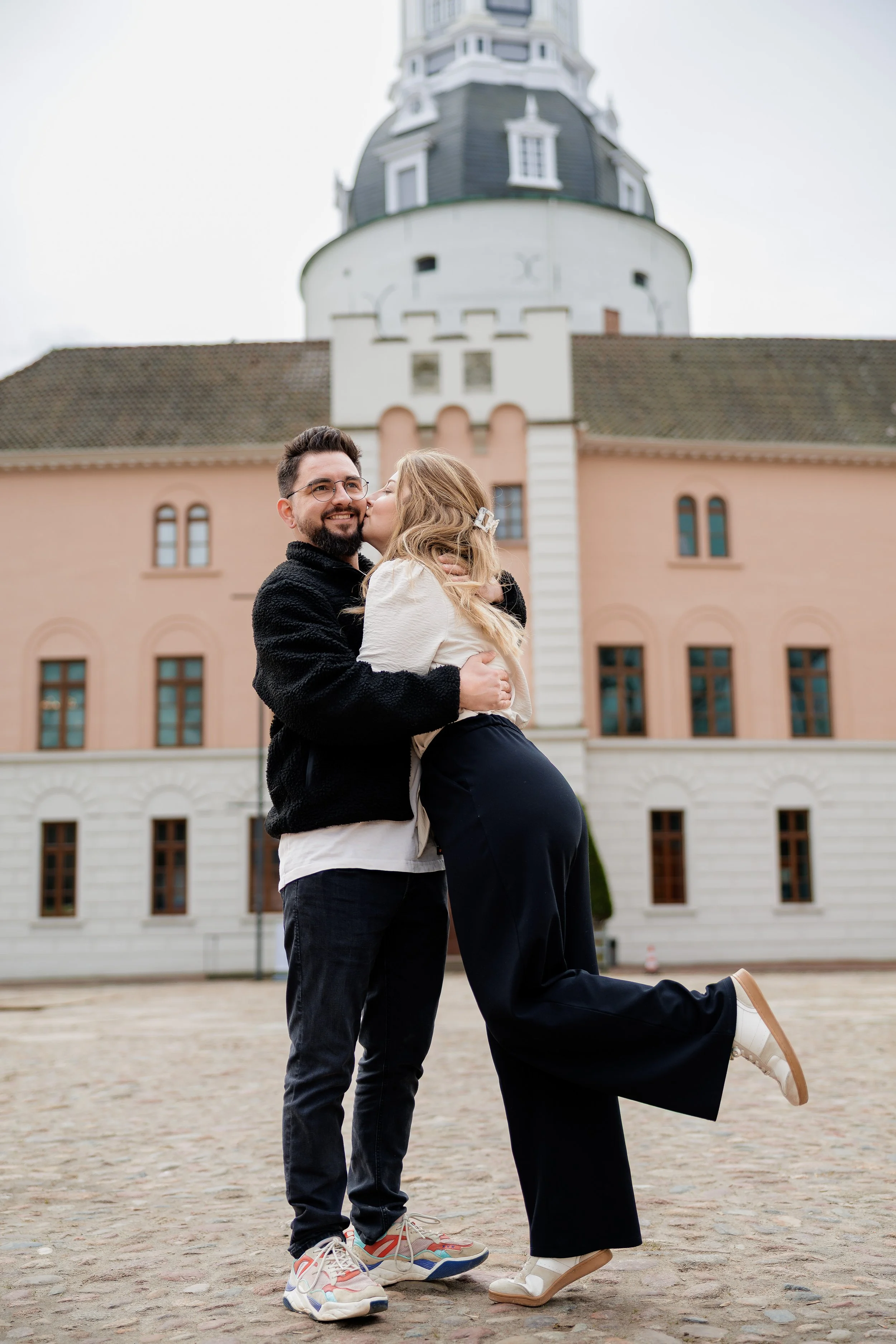 Ein lachendes Paar, ein Mann trägt eine Frau, während sie sich küssen, vor einem historischen Gebäude mit Türmen und Fenstern in Deutschland.