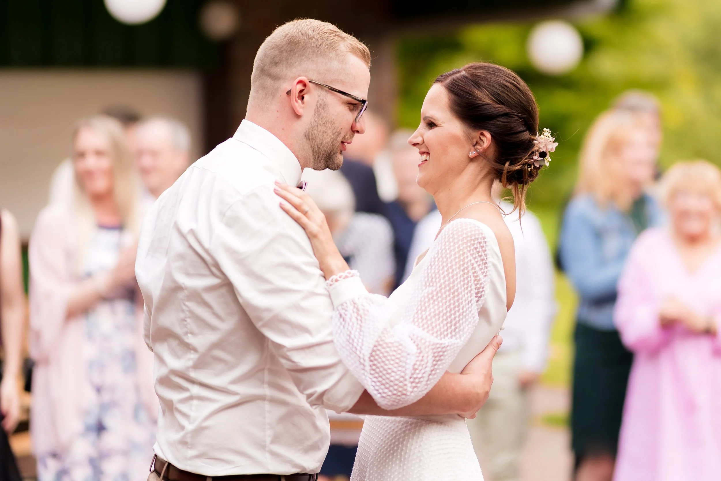 Ein Brautpaar tanzt bei einer Hochzeit im Freien, umgeben von Freunden und Familie, die im Hintergrund stehen und die Feier genießen.